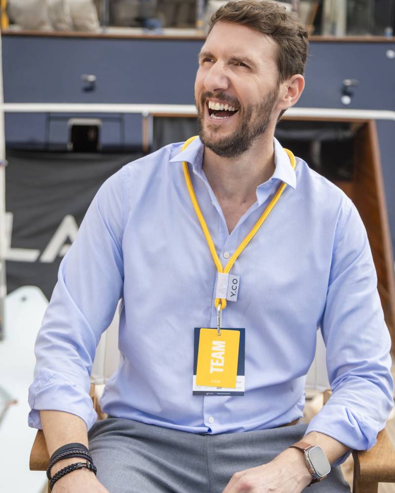 A laughing man wearing a light blue shirt and a yellow lanyard sits in the foreground against the backdrop of a luxury yacht.
