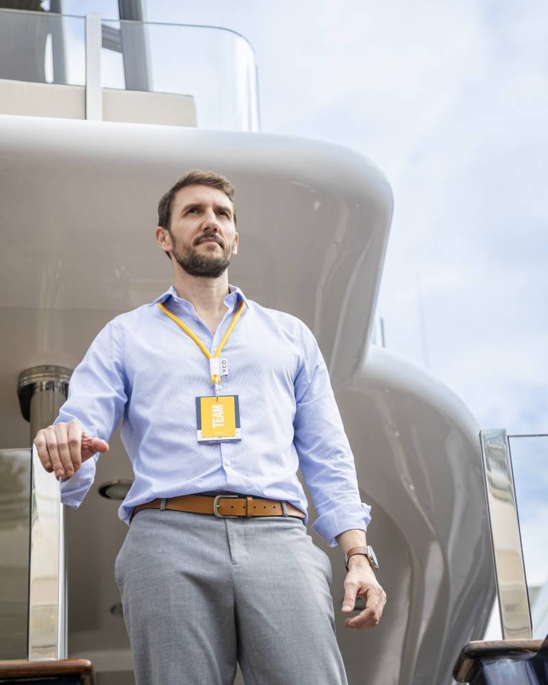 A man wearing a light blue shirt and grey trousers stands in the foreground against the backdrop of a sleek, white luxury yacht and a cloudy sky.