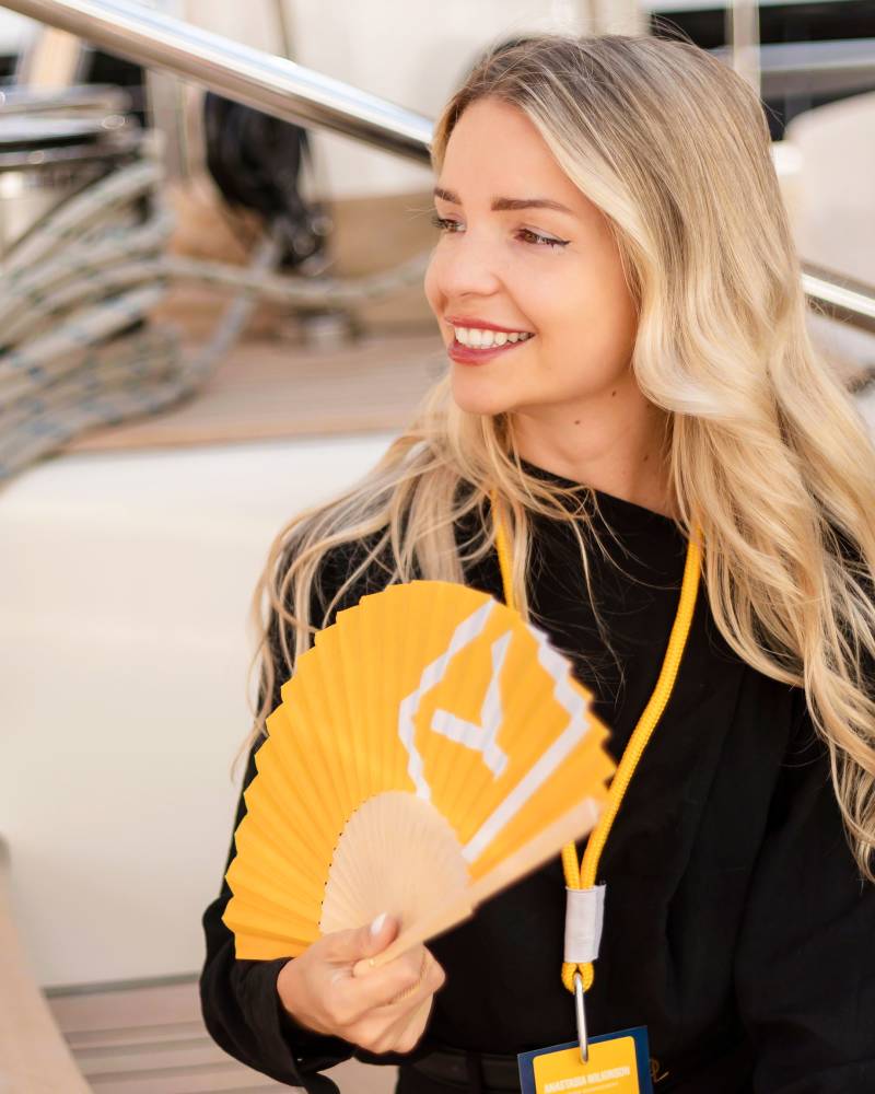 A smiling blonde woman in a black dress holds a yellow folding fan in the foreground, with the railing and deck of a boat blurred in the background.