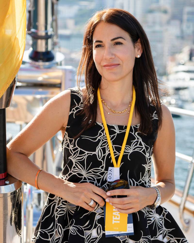 A woman in a black and white patterned dress stands on a boat with a blurred cityscape in the background.