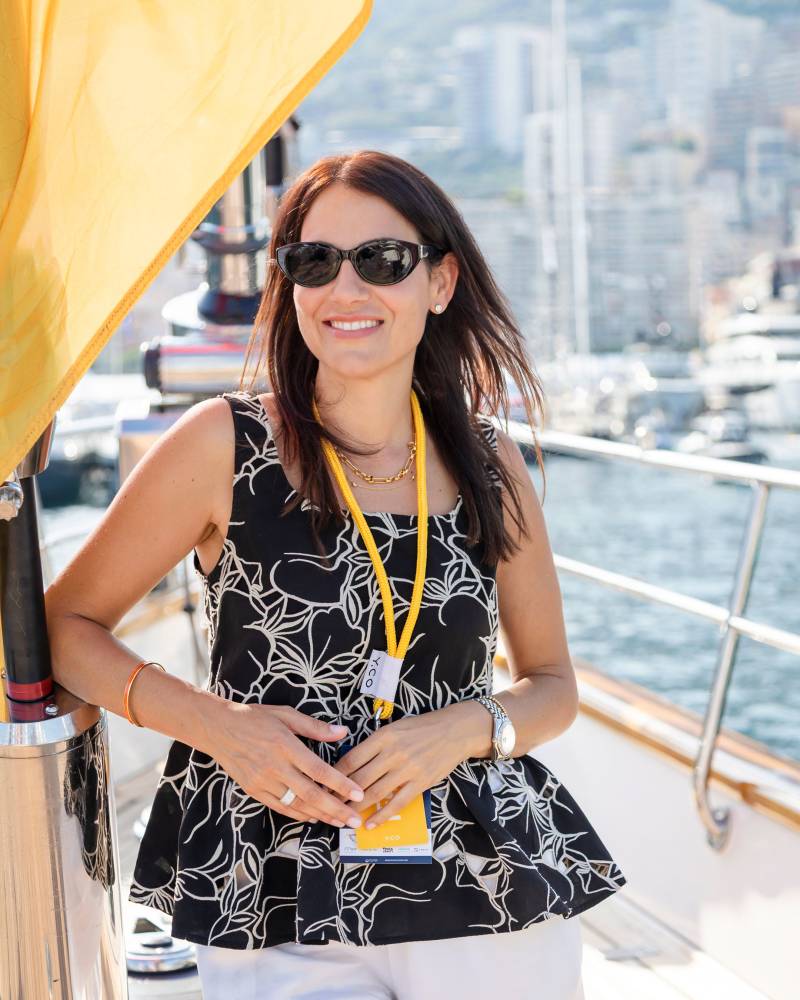 A woman wearing sunglasses and a patterned black top stands on a boat with a cityscape and harbor in the background.