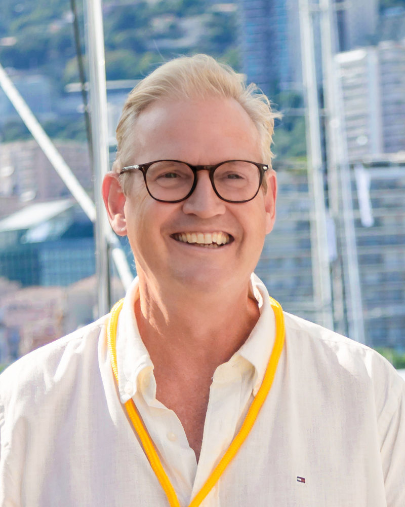A smiling man with glasses and a white shirt stands in the foreground against a blurred background of a harbor and city buildings.
