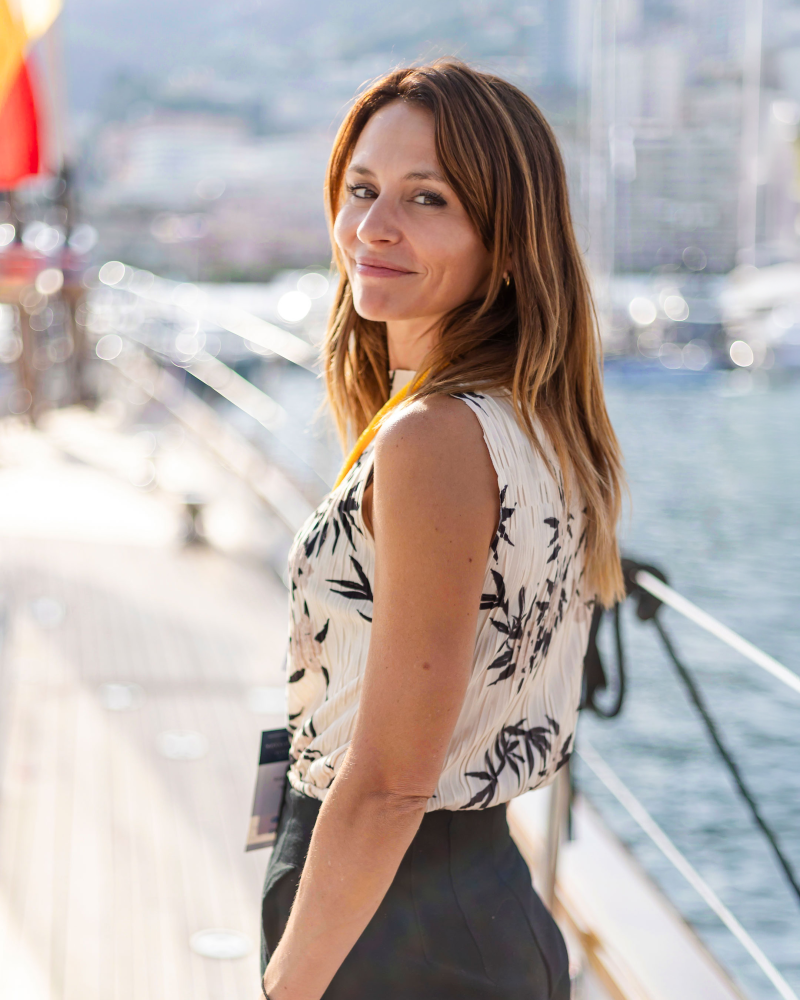 A woman in a patterned sleeveless top and dark trousers stands on a wooden boat deck with a blurred coastal city and harbor in the background.