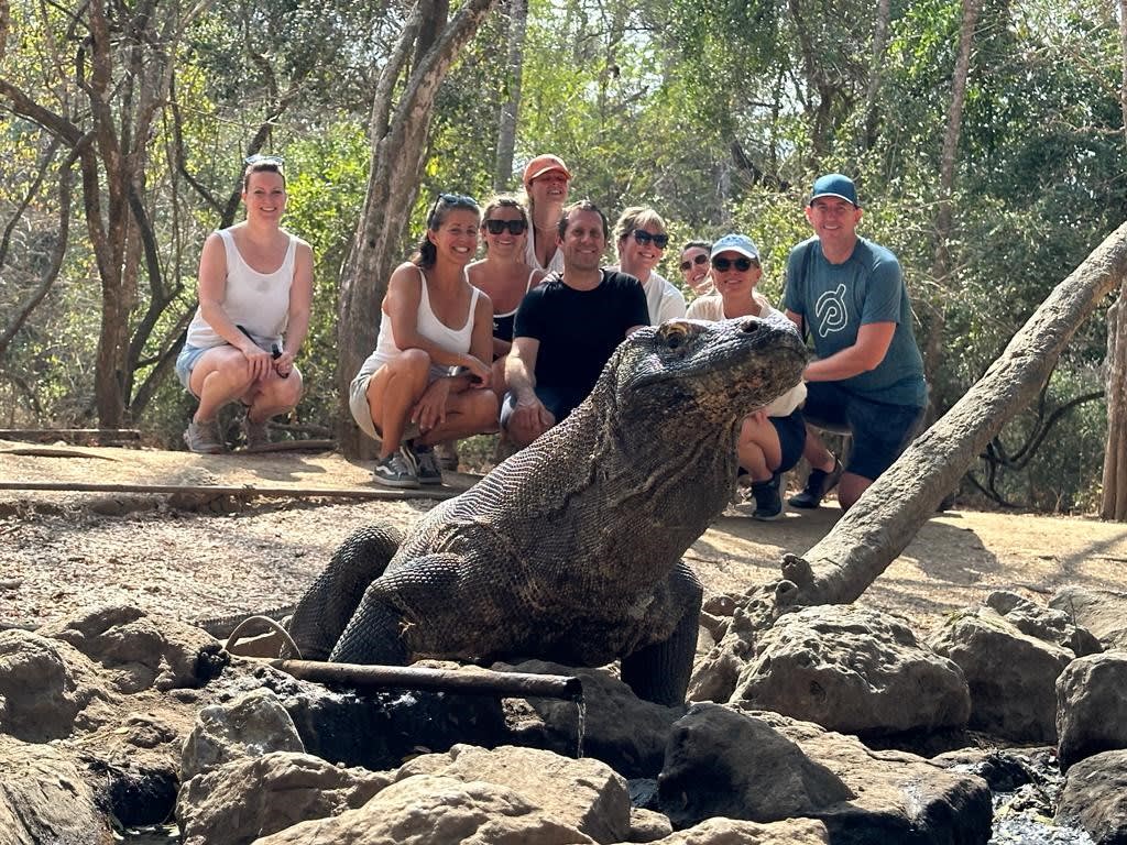A group of people, including several individuals wearing casual clothing, are gathered around a large lizard-like creature resting on a rocky surface in a wooded area.
