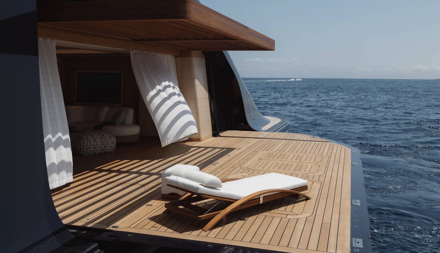 A white chaise lounge sits on a wooden deck in the foreground, with a luxurious interior visible through open curtains and the vast blue ocean stretching to the horizon in the background.