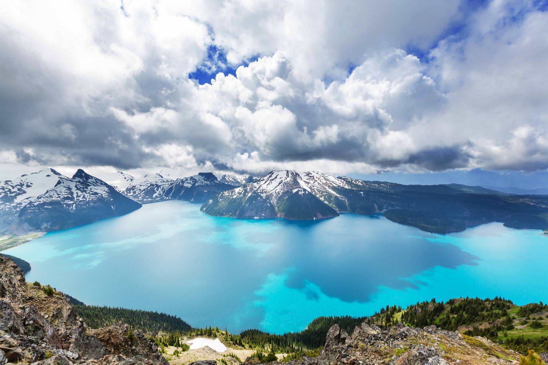 A vast turquoise lake is cradled by snow-capped mountains under a dramatic, cloudy sky.