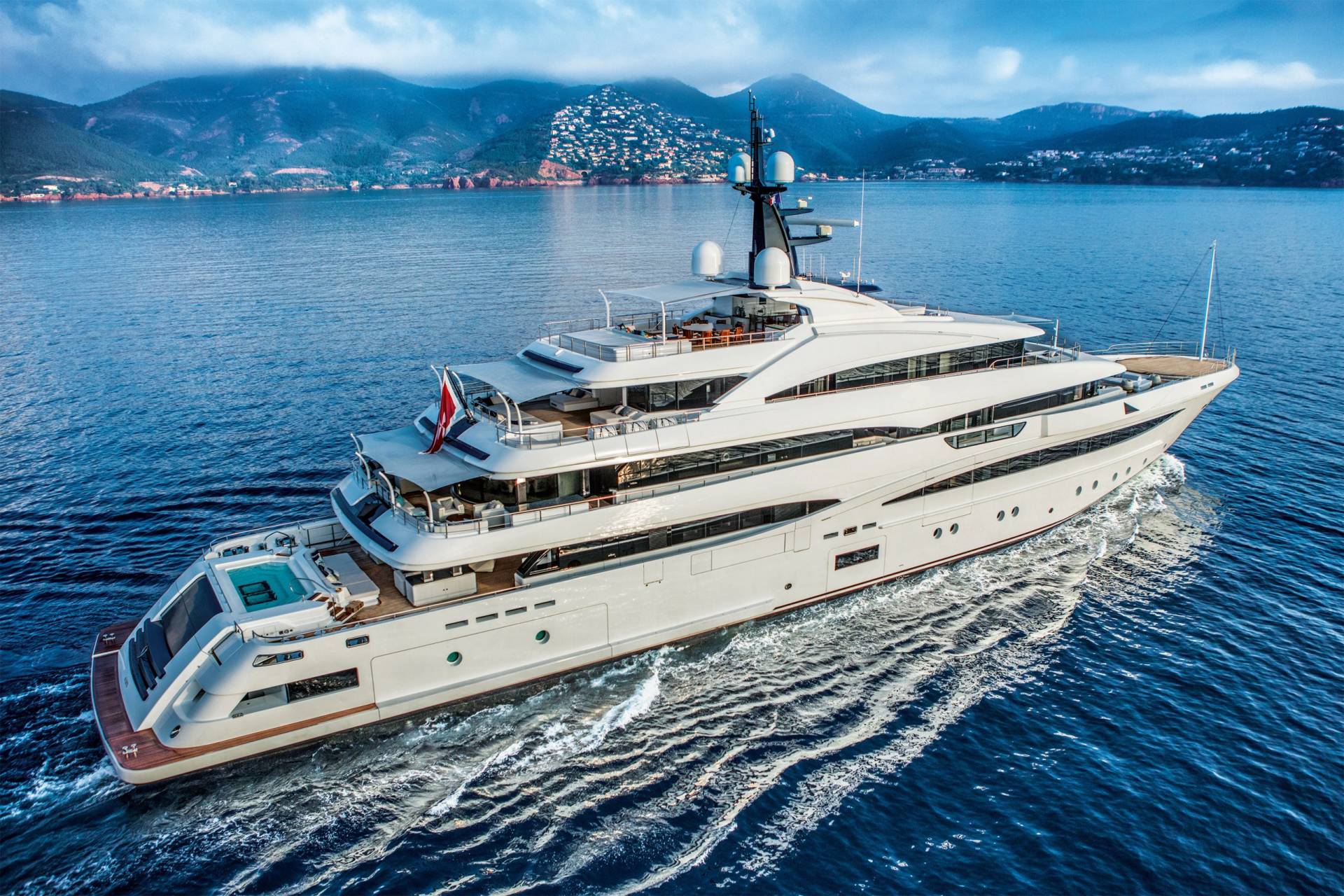 A luxurious white superyacht cruises through the deep blue sea, with a picturesque coastline and mountains in the background.