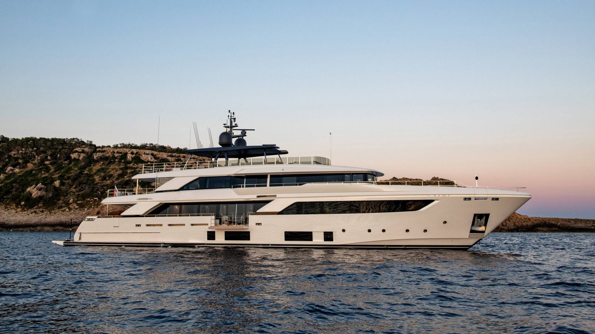 A sleek white yacht cruises on the calm blue sea near a rocky, tree-covered coastline at sunset.