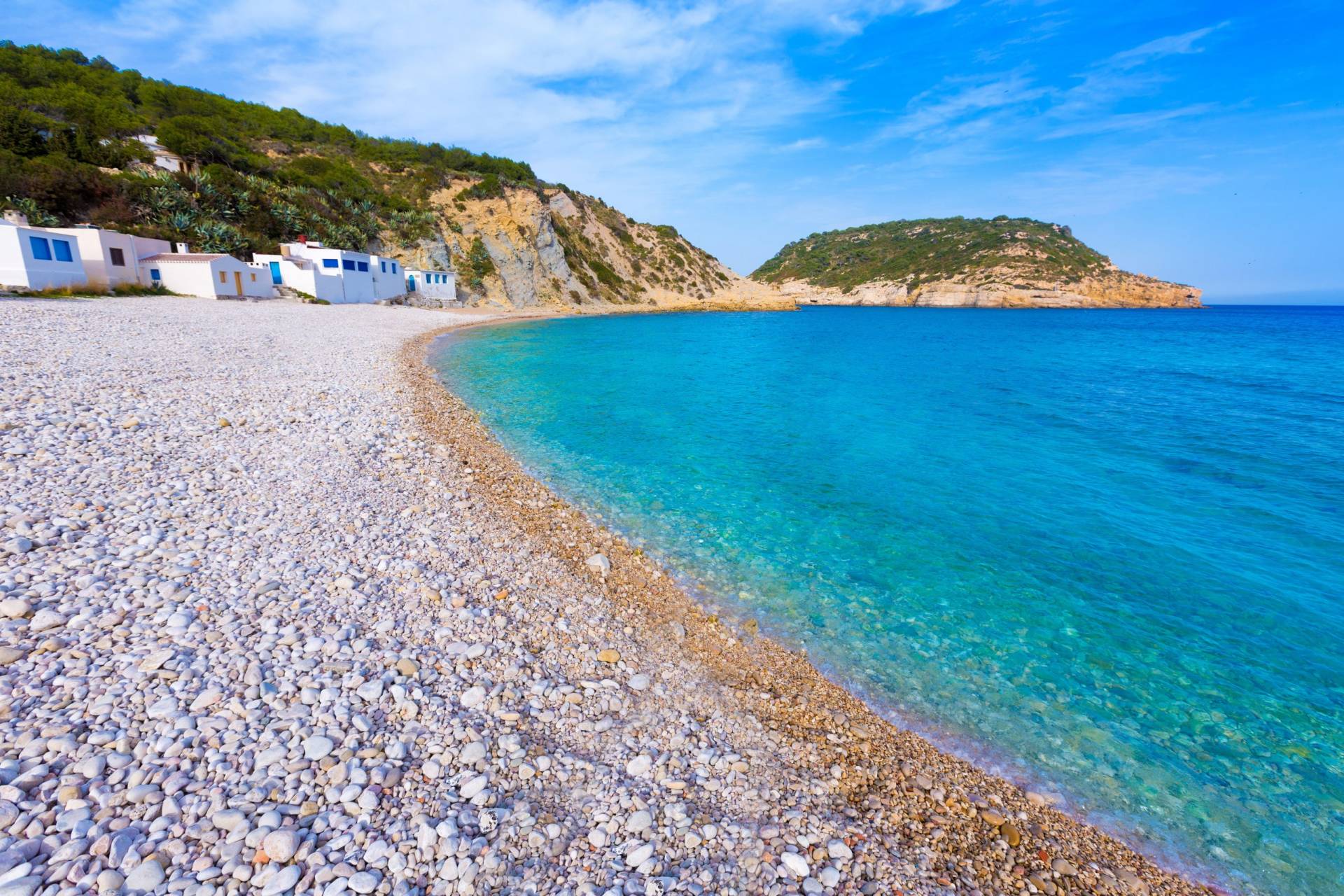 White buildings line a pebble beach leading to the crystal-clear turquoise waters of a secluded cove.