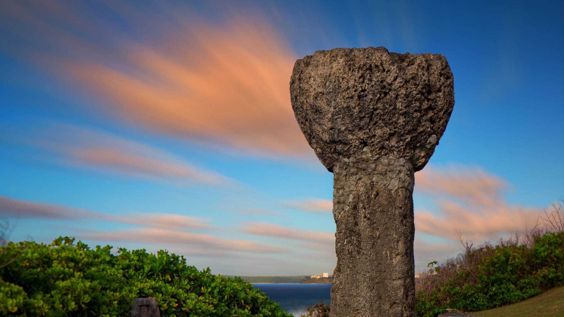 A weathered stone monument stands against a vibrant sky streaked with pink clouds over the ocean.