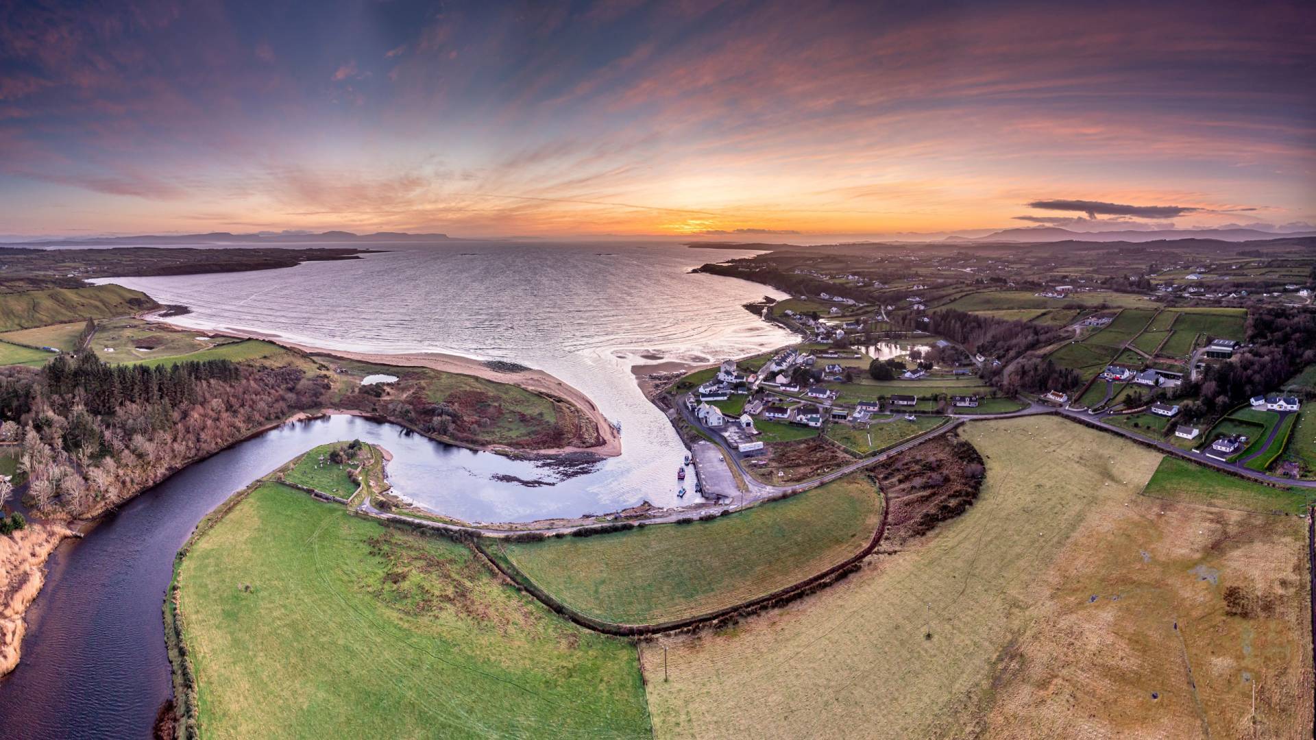 A breathtaking panoramic view captures a coastal village nestled beside a tranquil river estuary at sunset.