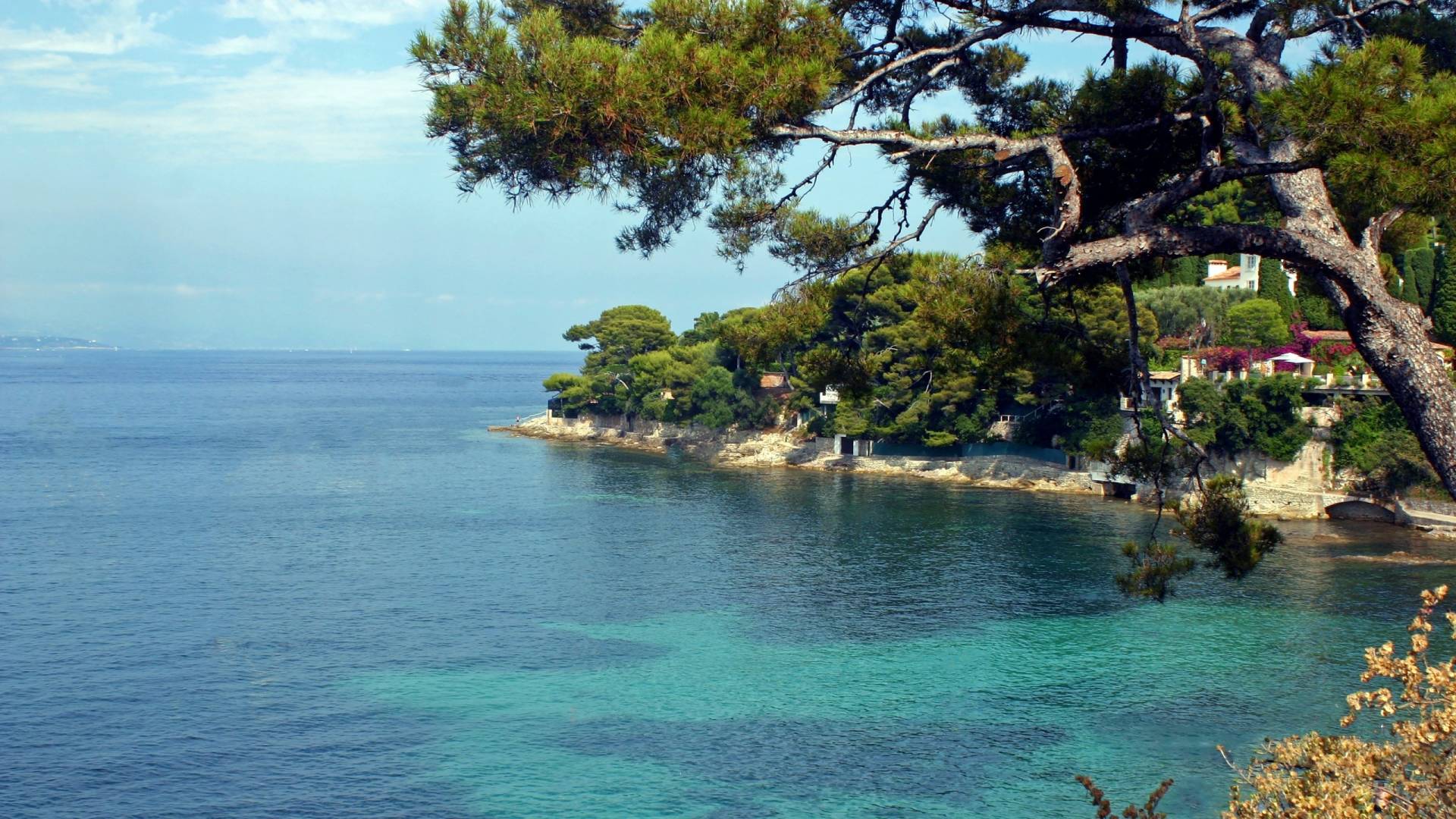 Crystal-clear turquoise waters lap against a rocky coastline dotted with lush green trees and charming villas.