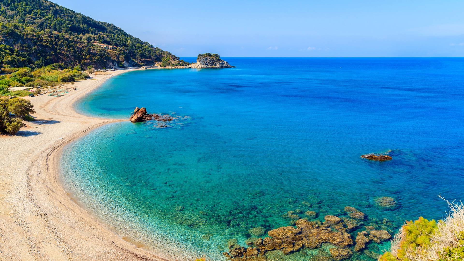 A crescent-shaped beach with turquoise waters meets a lush green hillside under a clear blue sky.