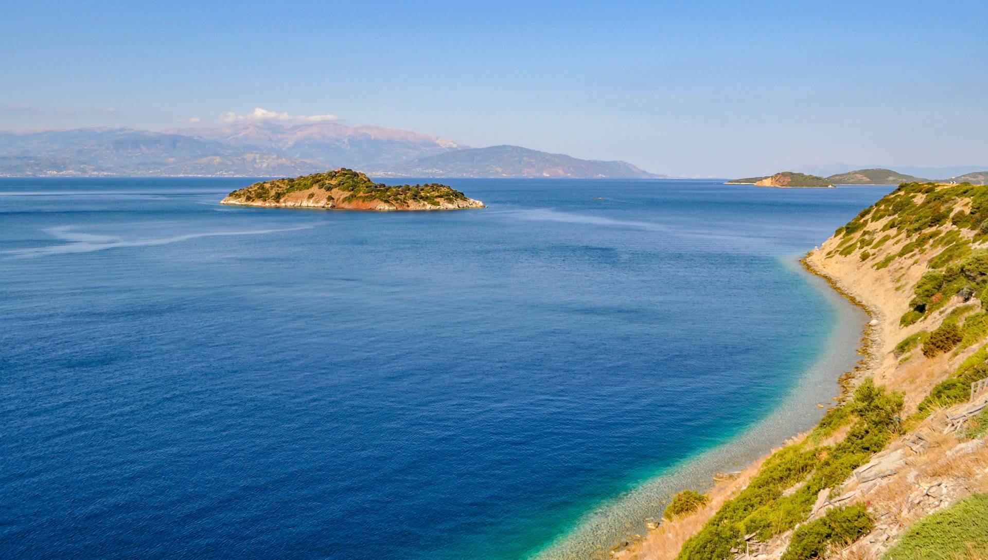 A small, tree-covered island sits in the vast blue sea under a clear sky, with a rugged coastline in the foreground.