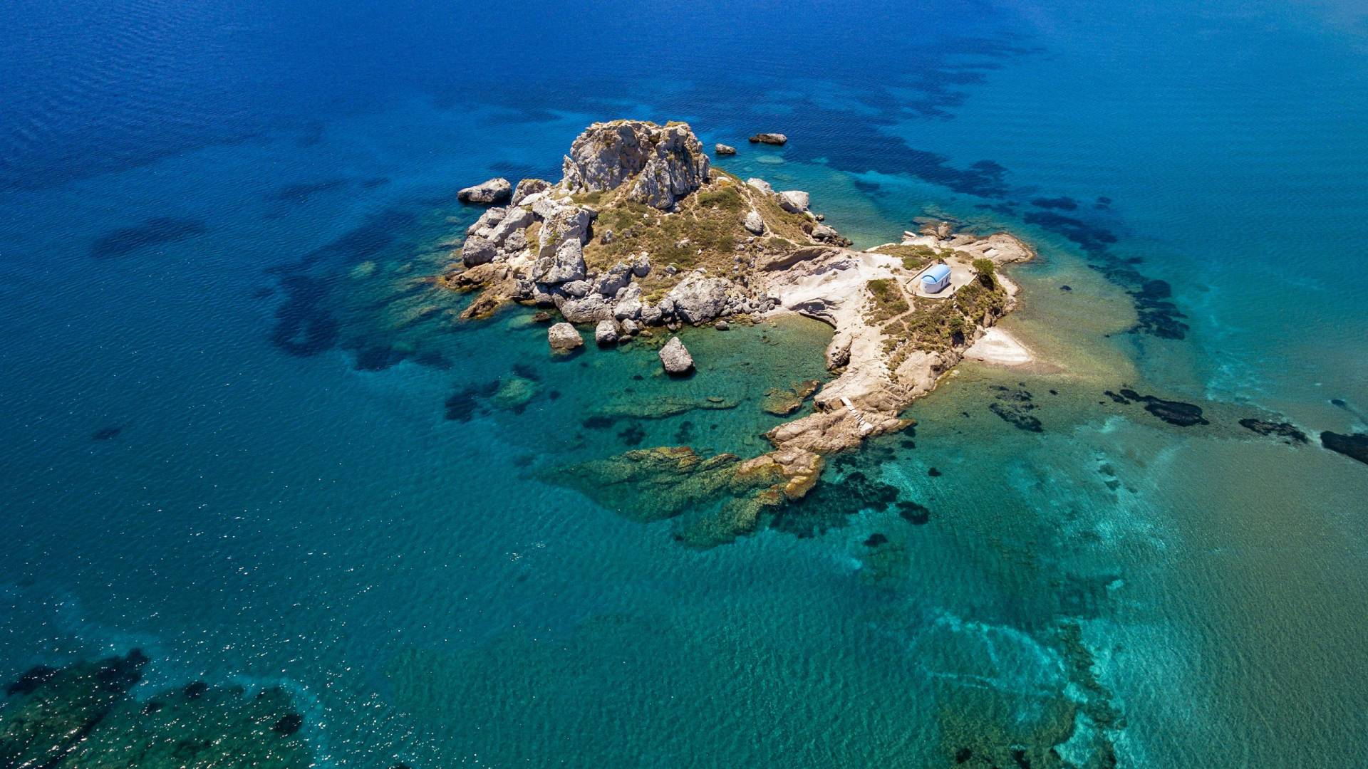 A small white church with a blue dome stands on a rocky island surrounded by crystal-clear turquoise waters.