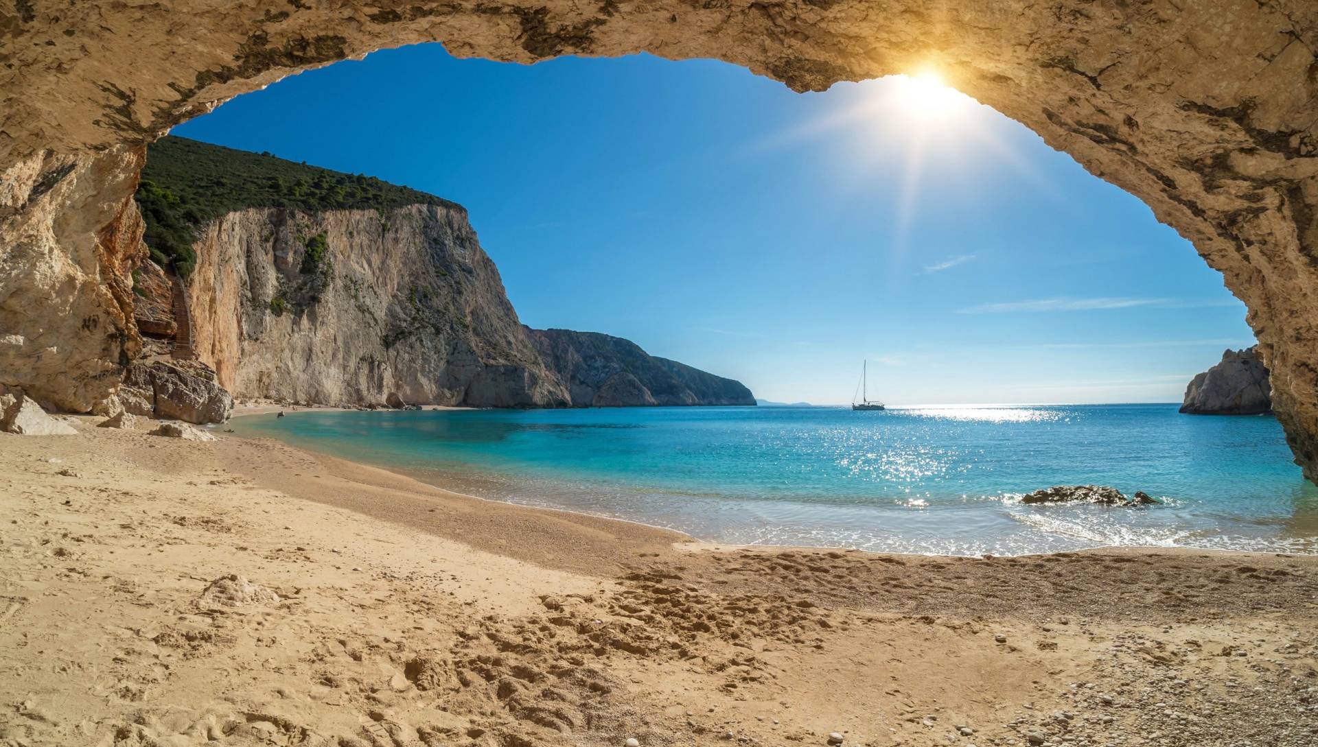 Sunlight streams through a cave opening onto a pristine beach with turquoise waters and a distant sailboat.