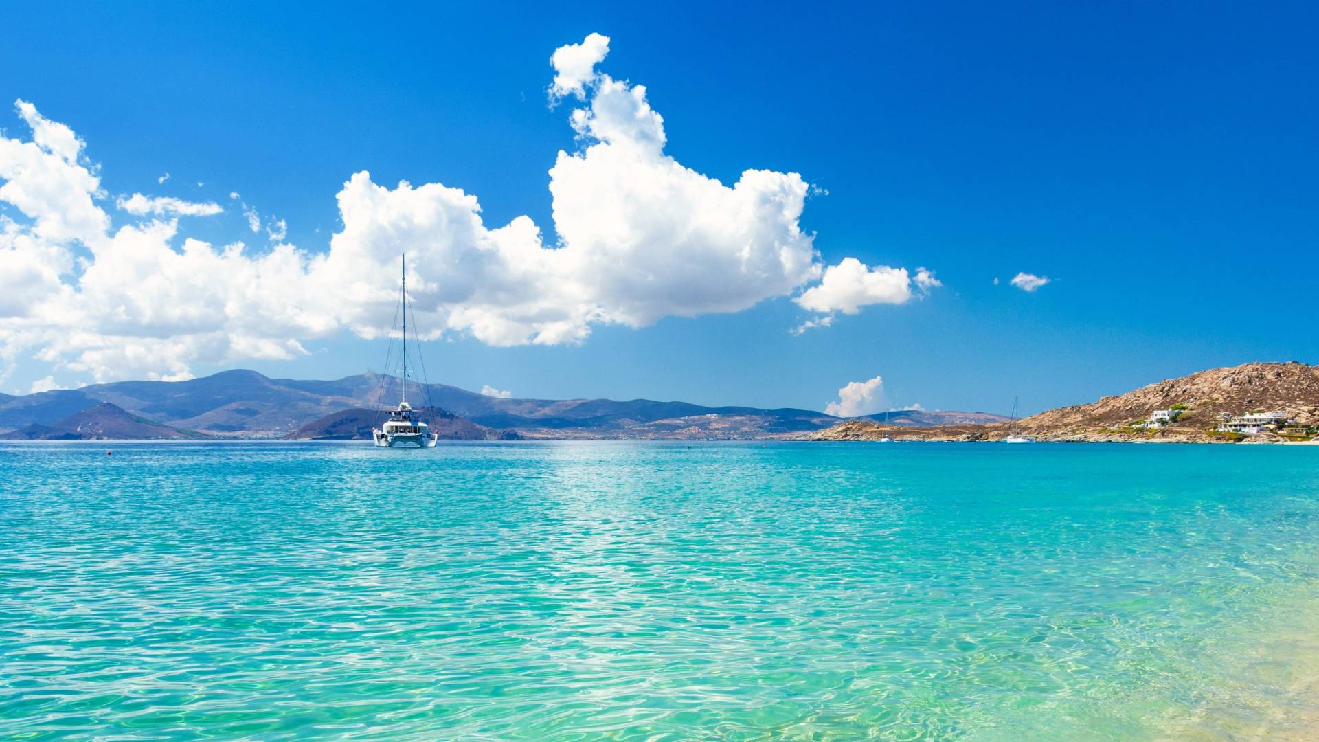 A lone sailboat glides across the turquoise waters of a Greek island under a bright blue sky.