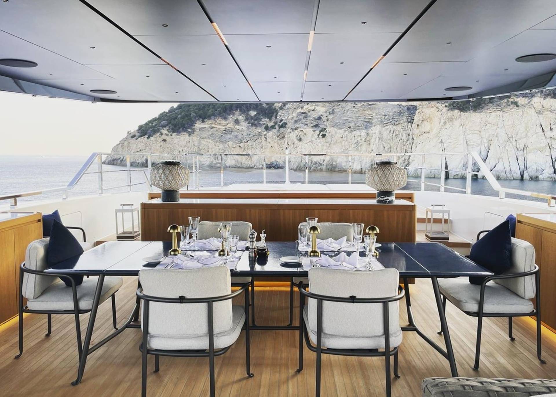 An elegant outdoor dining setup on a yacht offers a breathtaking view of a rocky coastline.