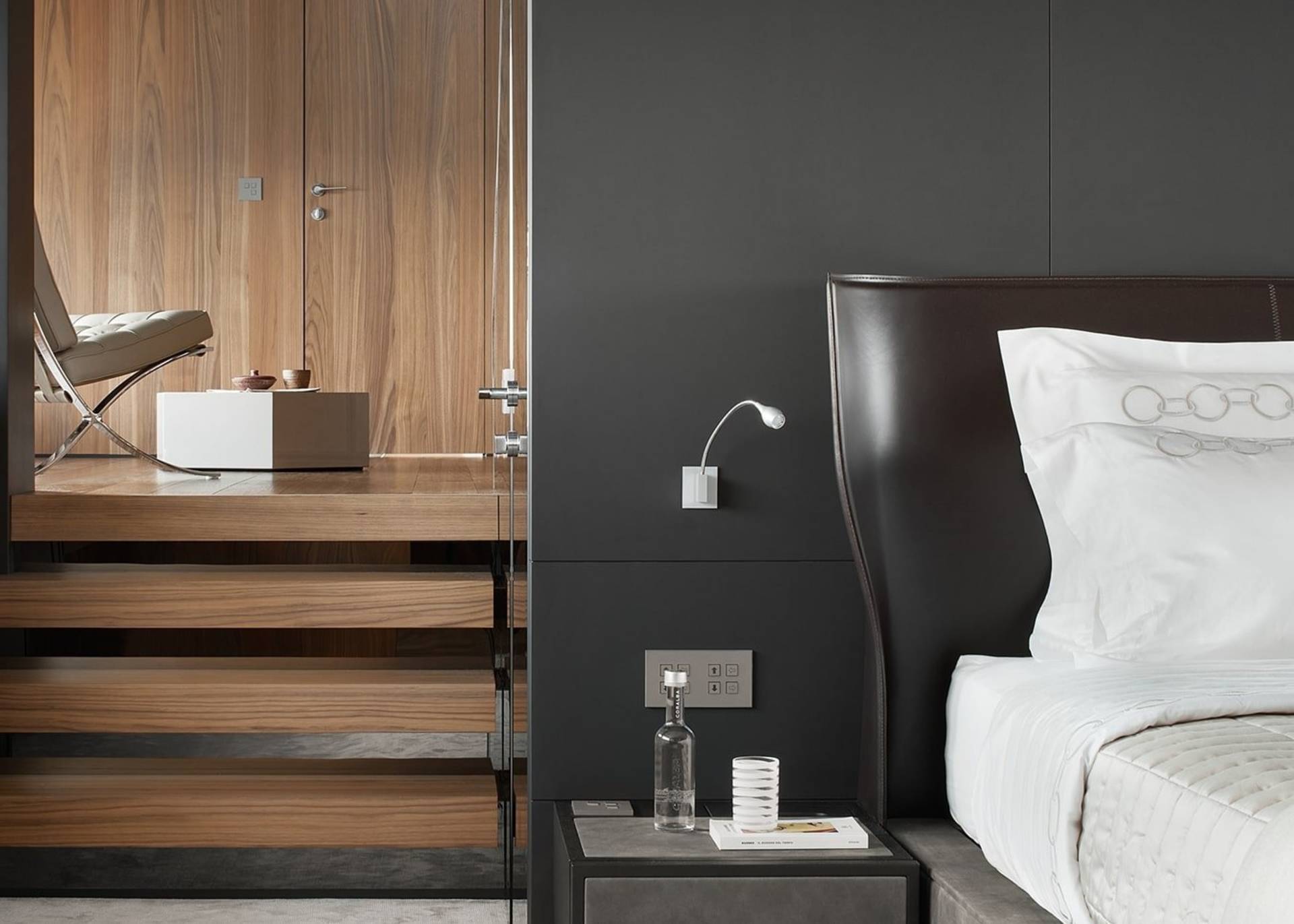 A modern bedroom features a sleek leather headboard, a minimalist bedside table with a glass of water, and a wooden staircase leading to a sitting area.