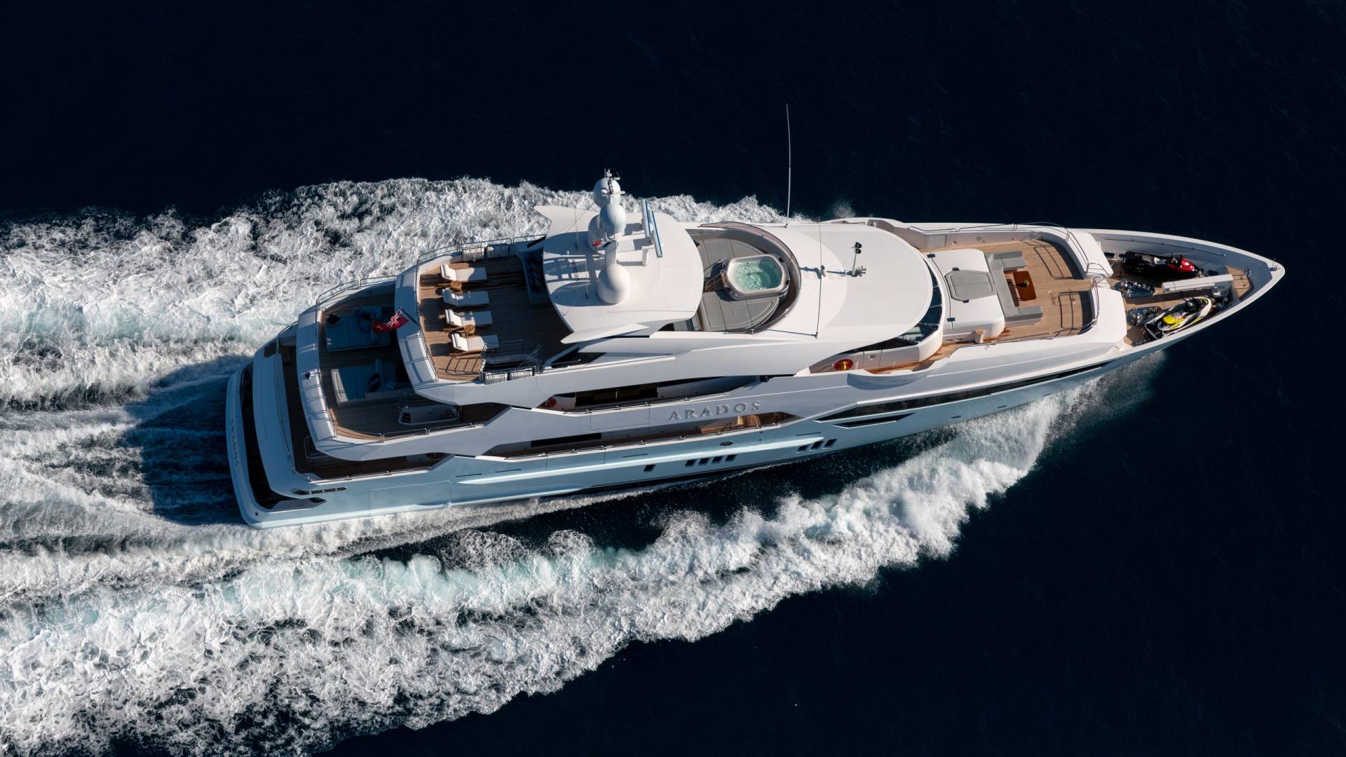 A luxurious yacht named Arados glides through the deep blue ocean, leaving a trail of white foam in its wake.