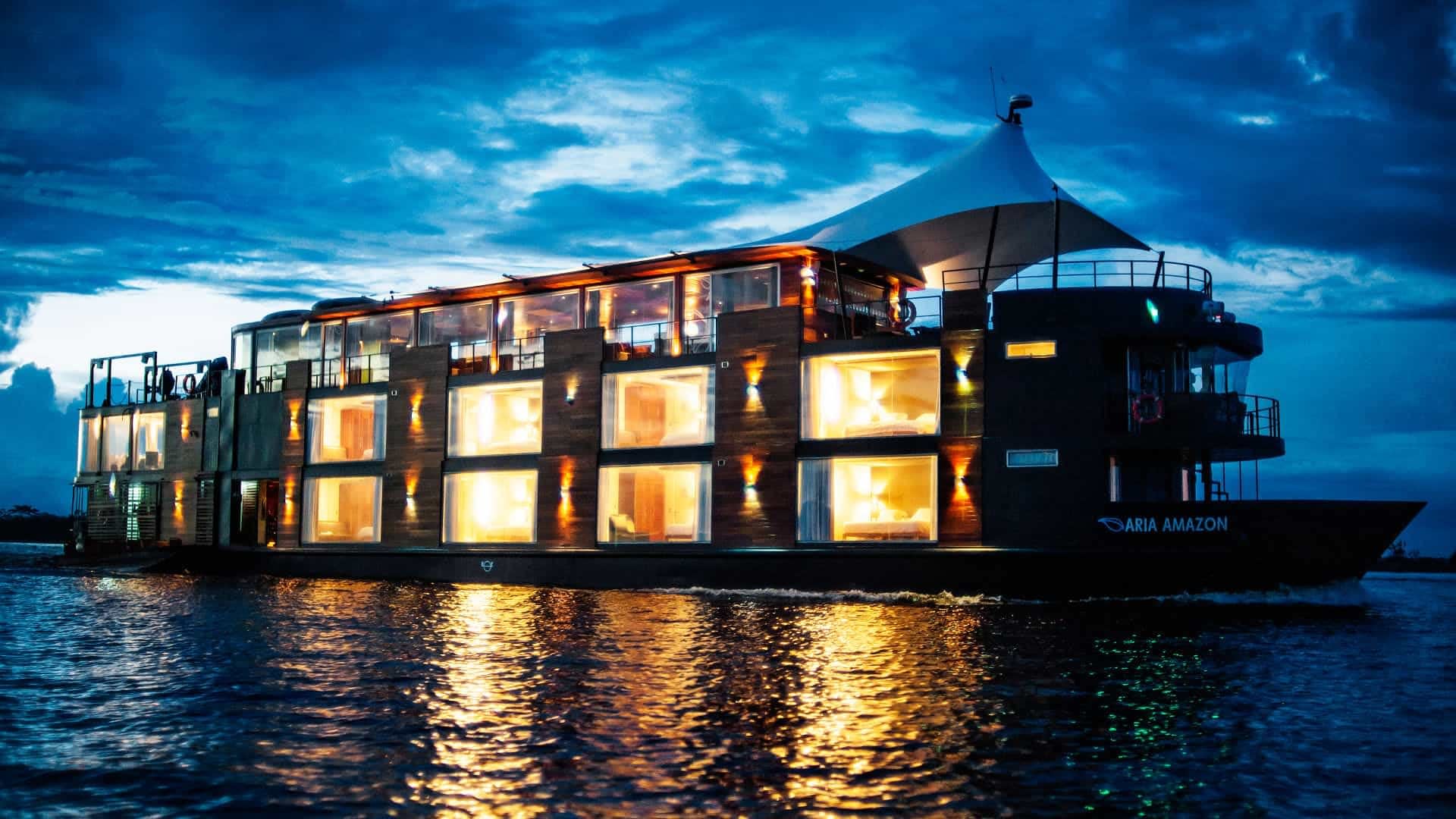 The Aria Amazon luxury riverboat glides through dark waters under a dramatic twilight sky.