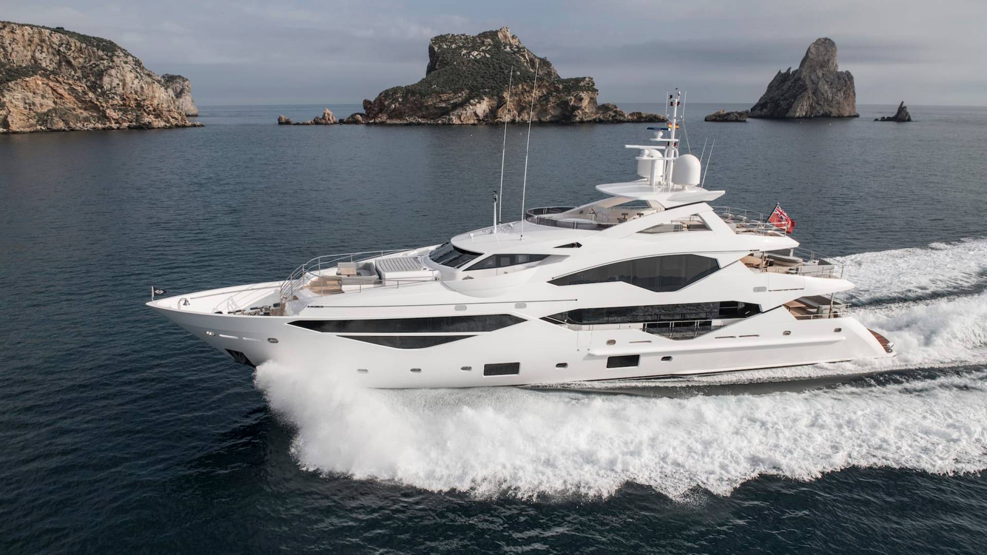 A sleek white yacht glides through the dark blue ocean, leaving a foamy wake against a backdrop of rugged islands.