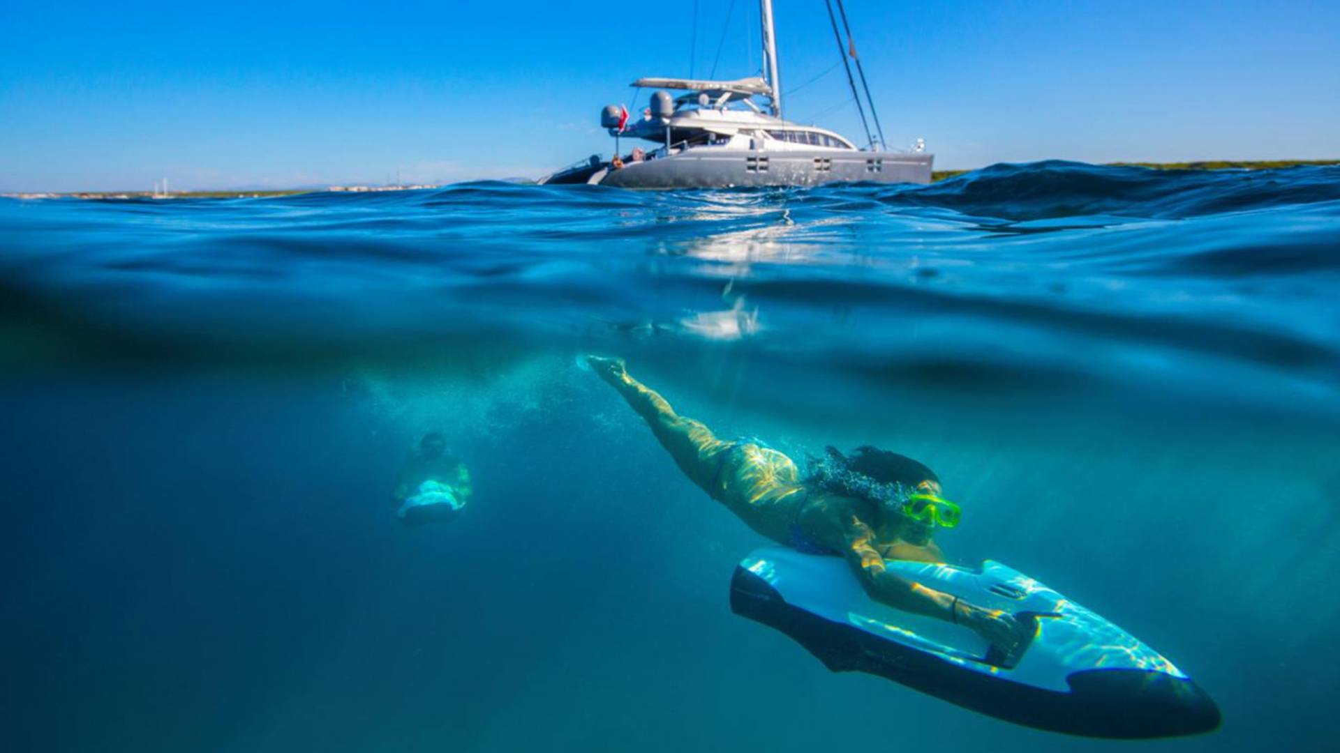 A woman glides through clear blue water on a sea scooter, with a luxury catamaran anchored nearby.