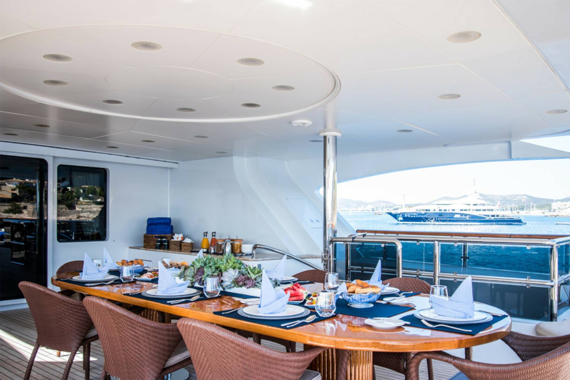 A beautifully set table awaits on a yacht deck, offering a picturesque view of the sea and a distant luxury vessel.