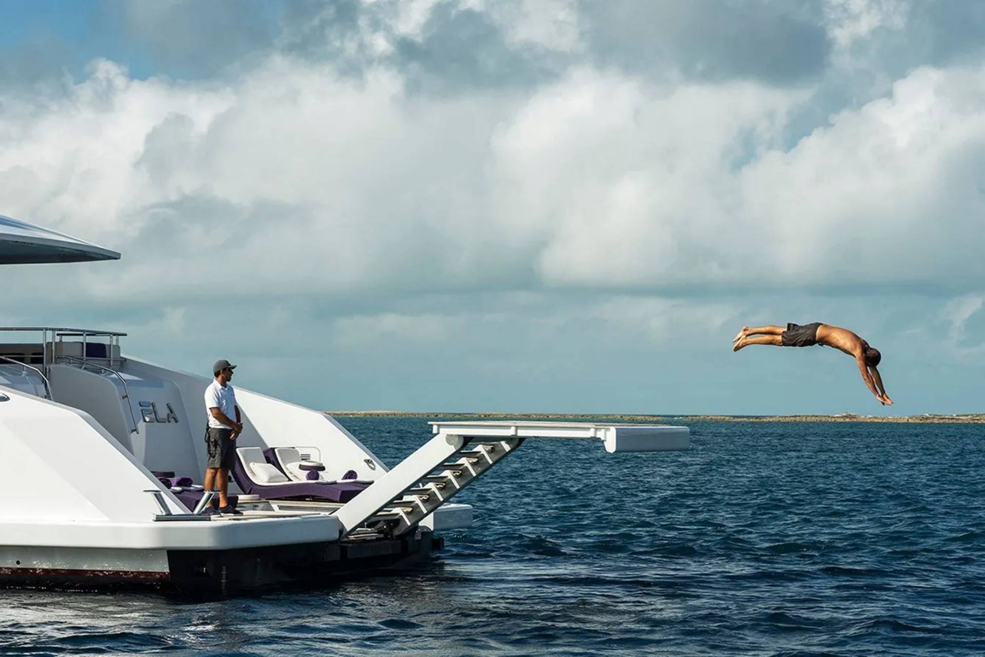 A man dives from a luxury yacht into the clear blue ocean under a cloudy sky.