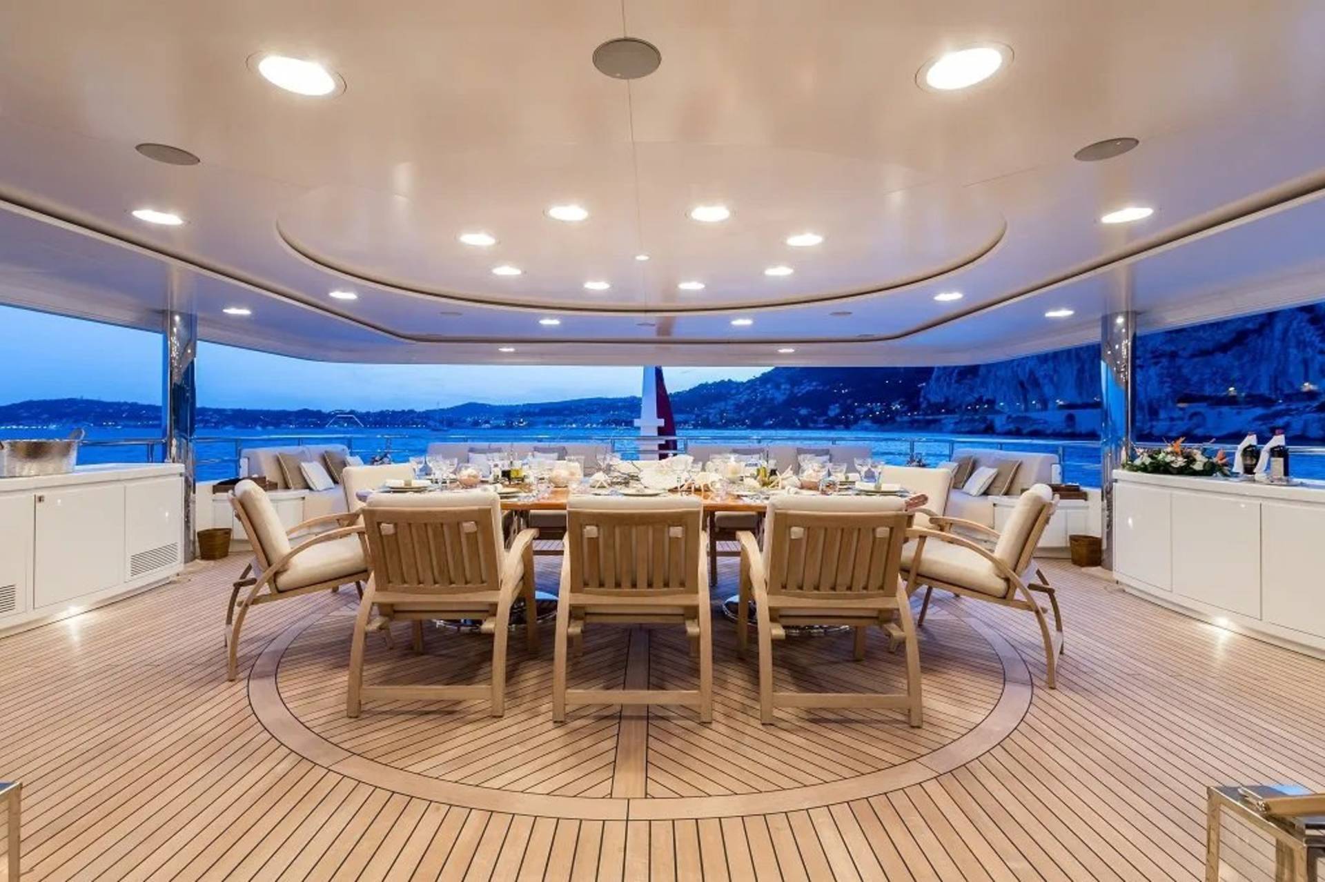A beautifully set dining table awaits guests on the deck of a luxurious yacht, overlooking a picturesque coastal landscape at dusk.