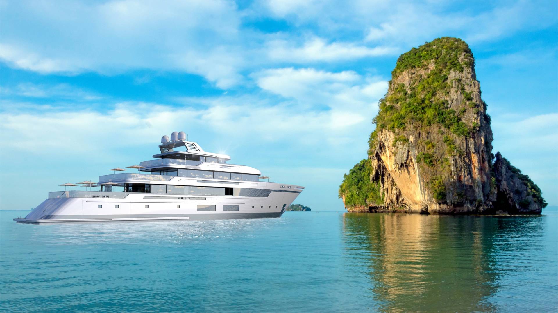 A luxurious modern yacht cruises past a majestic, verdant limestone karst island under a bright blue sky.