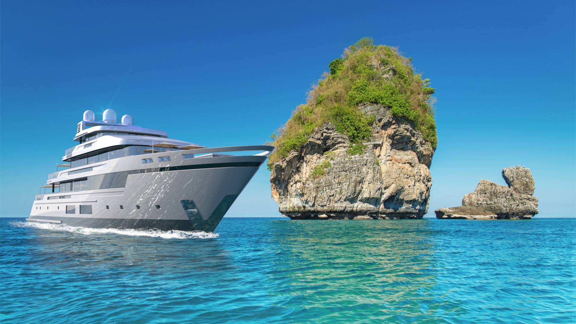 A sleek modern yacht glides through turquoise waters past a lush, rocky island under a clear blue sky.