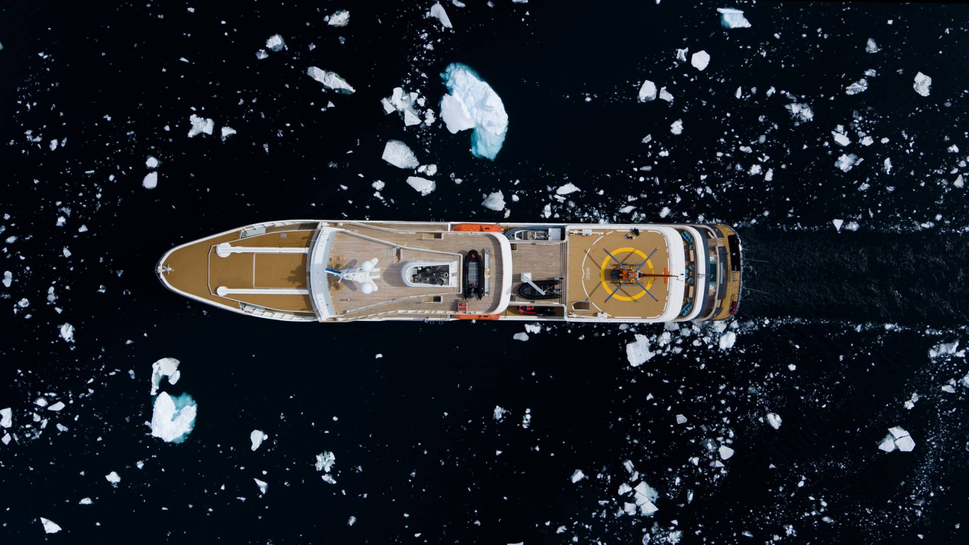 An aerial view captures a luxury yacht navigating through a dark, icy sea dotted with small ice floes.