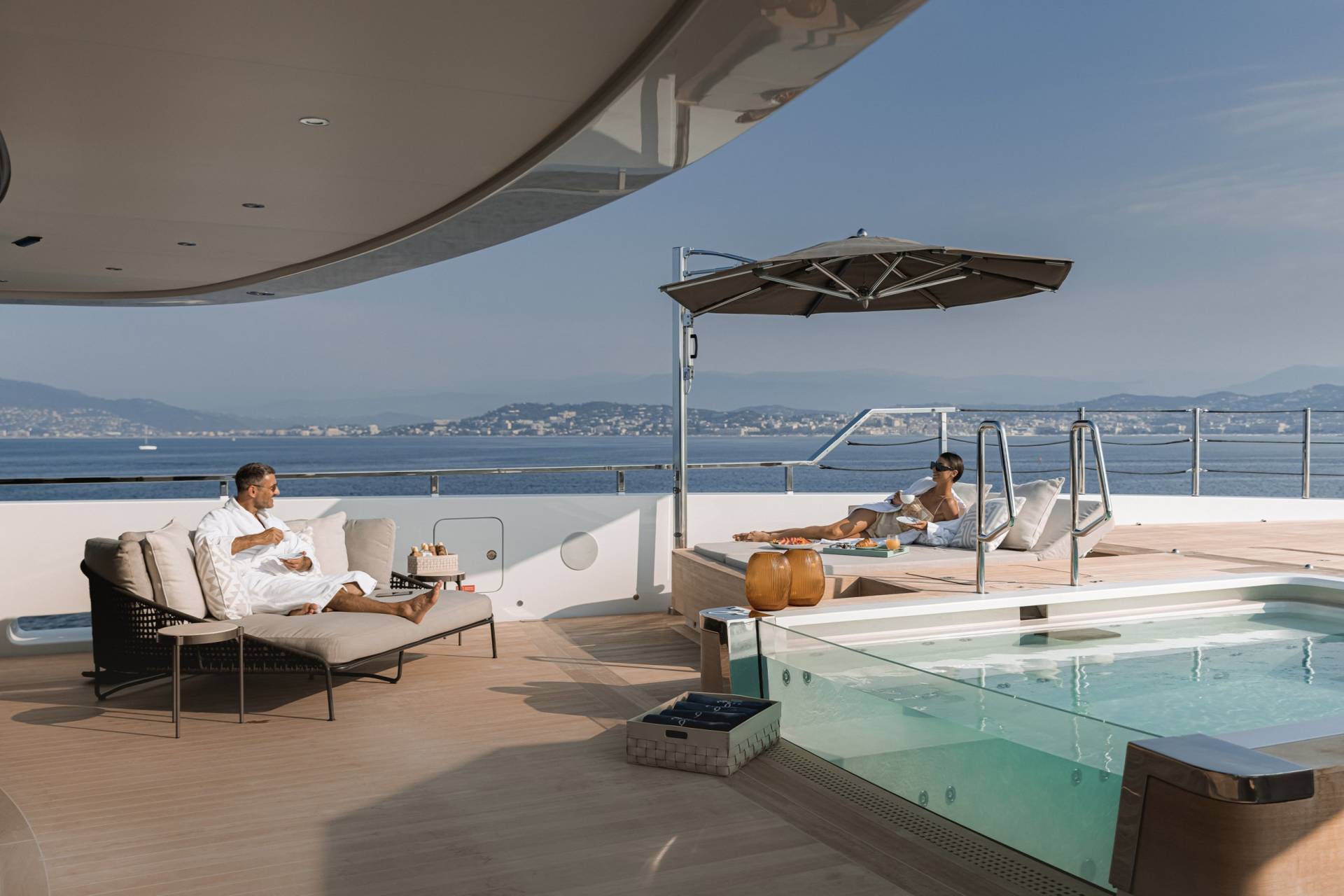 A man in a white robe sips coffee while a woman relaxes on a sun lounger by a glass-walled hot tub on a luxury yacht.