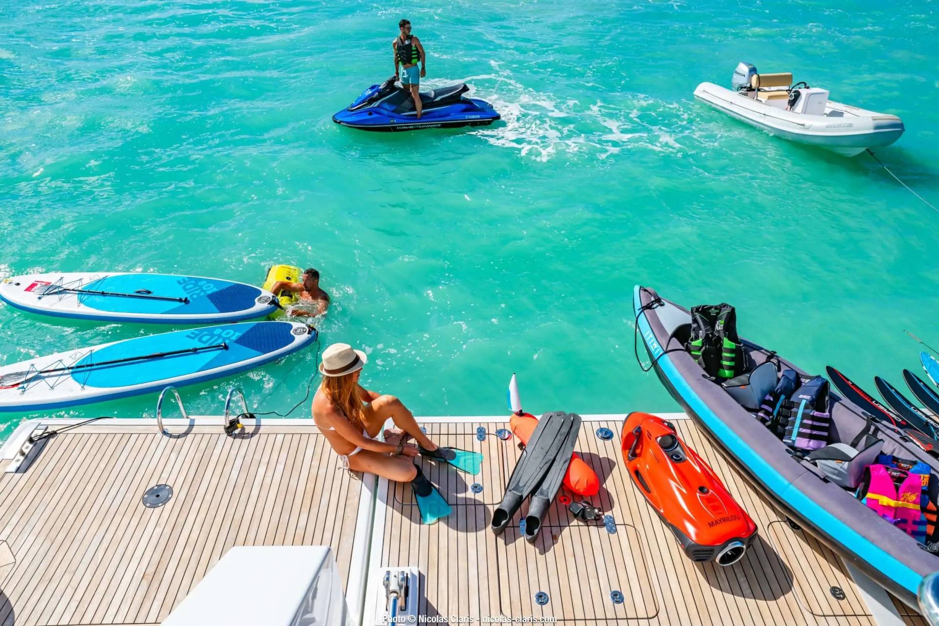 A woman in a hat relaxes on a yacht deck surrounded by water sports equipment and turquoise waters.