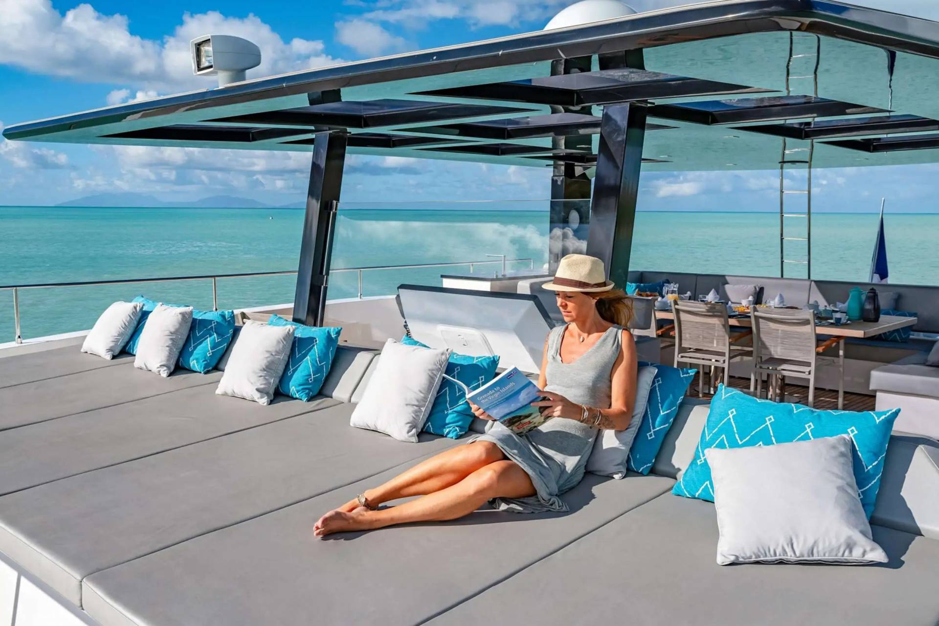A woman in a straw hat relaxes on a yacht's sundeck, reading a book with the turquoise ocean stretching out before her.