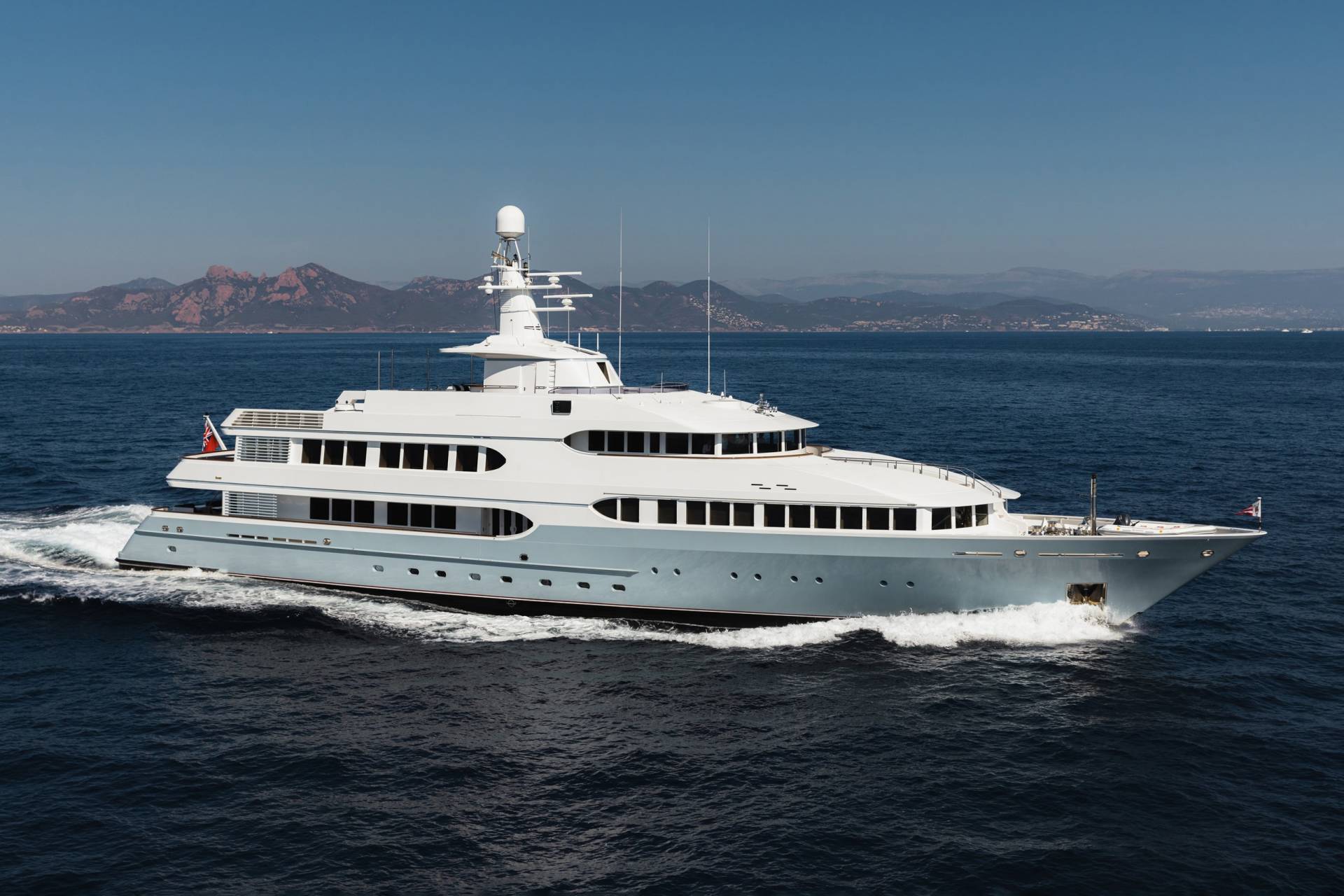A luxurious superyacht glides through the deep blue sea with a mountainous coastline in the distance.