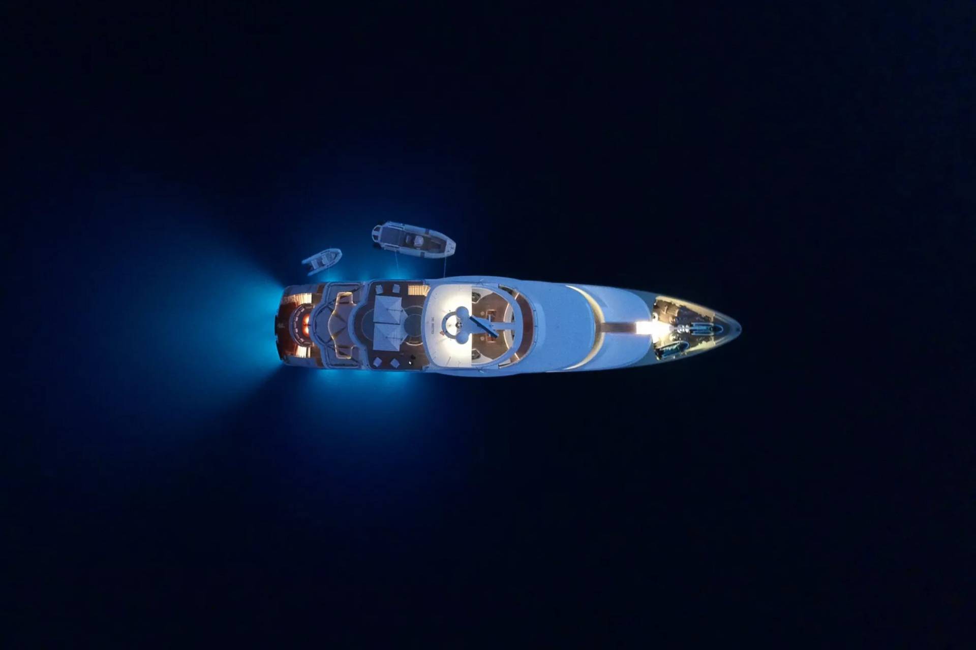 A luxury yacht glows with underwater lights as two tenders bob nearby in the dark ocean.