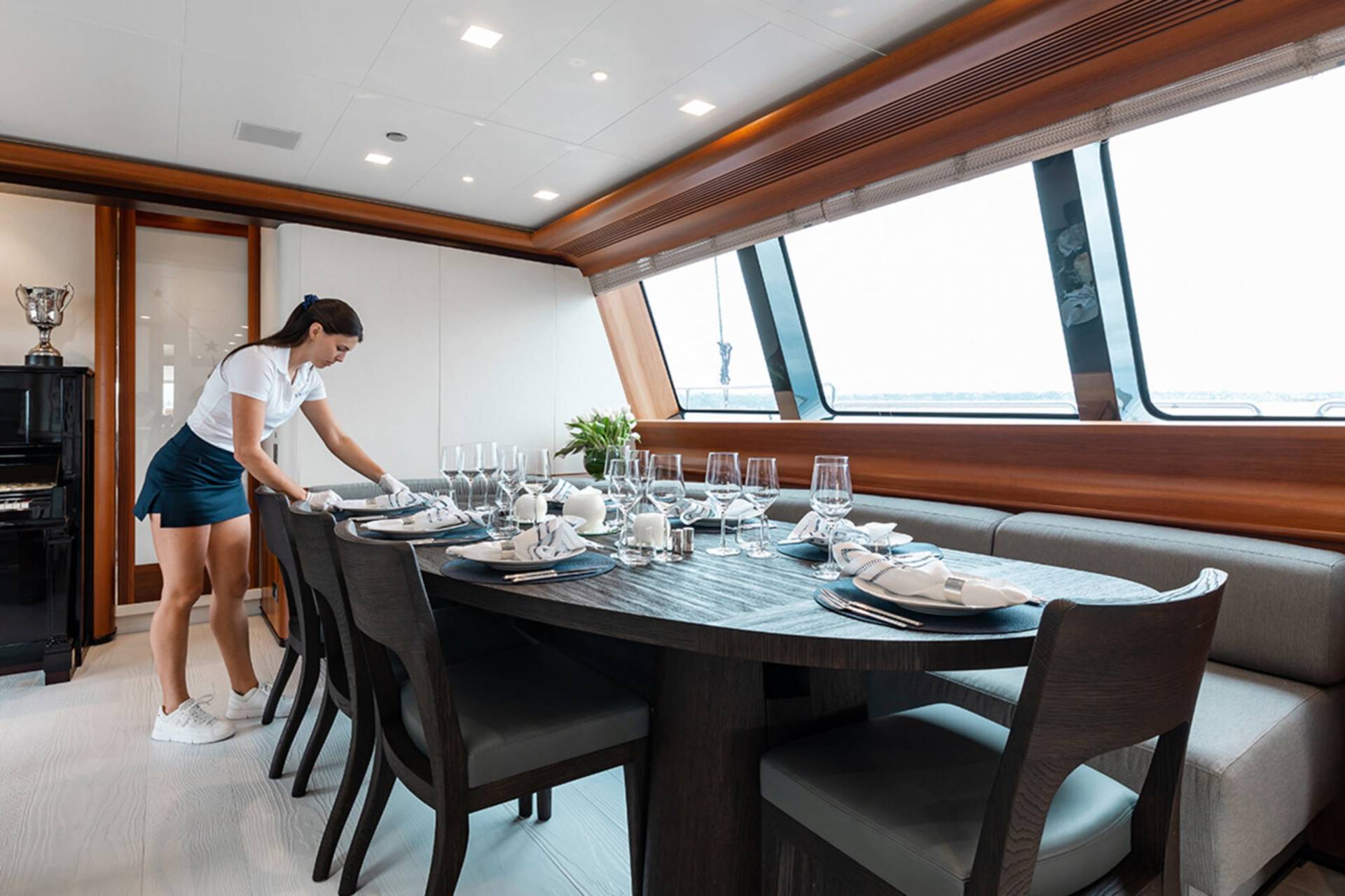 A woman in a white polo and blue skirt sets a formal dining table on a yacht with a panoramic view.