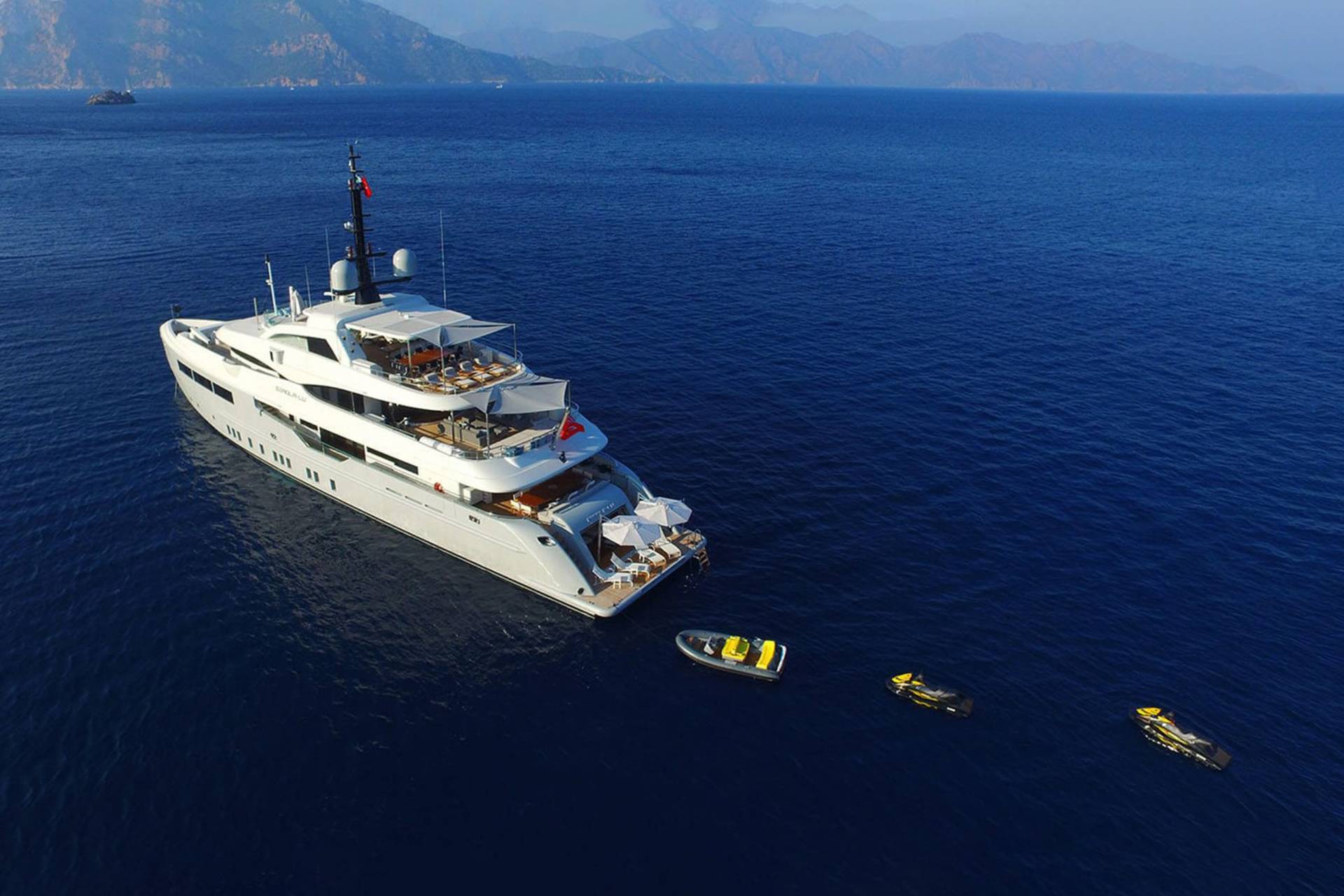 A luxurious white yacht cruises through deep blue waters near a mountainous coastline, accompanied by a tender and jet skis.