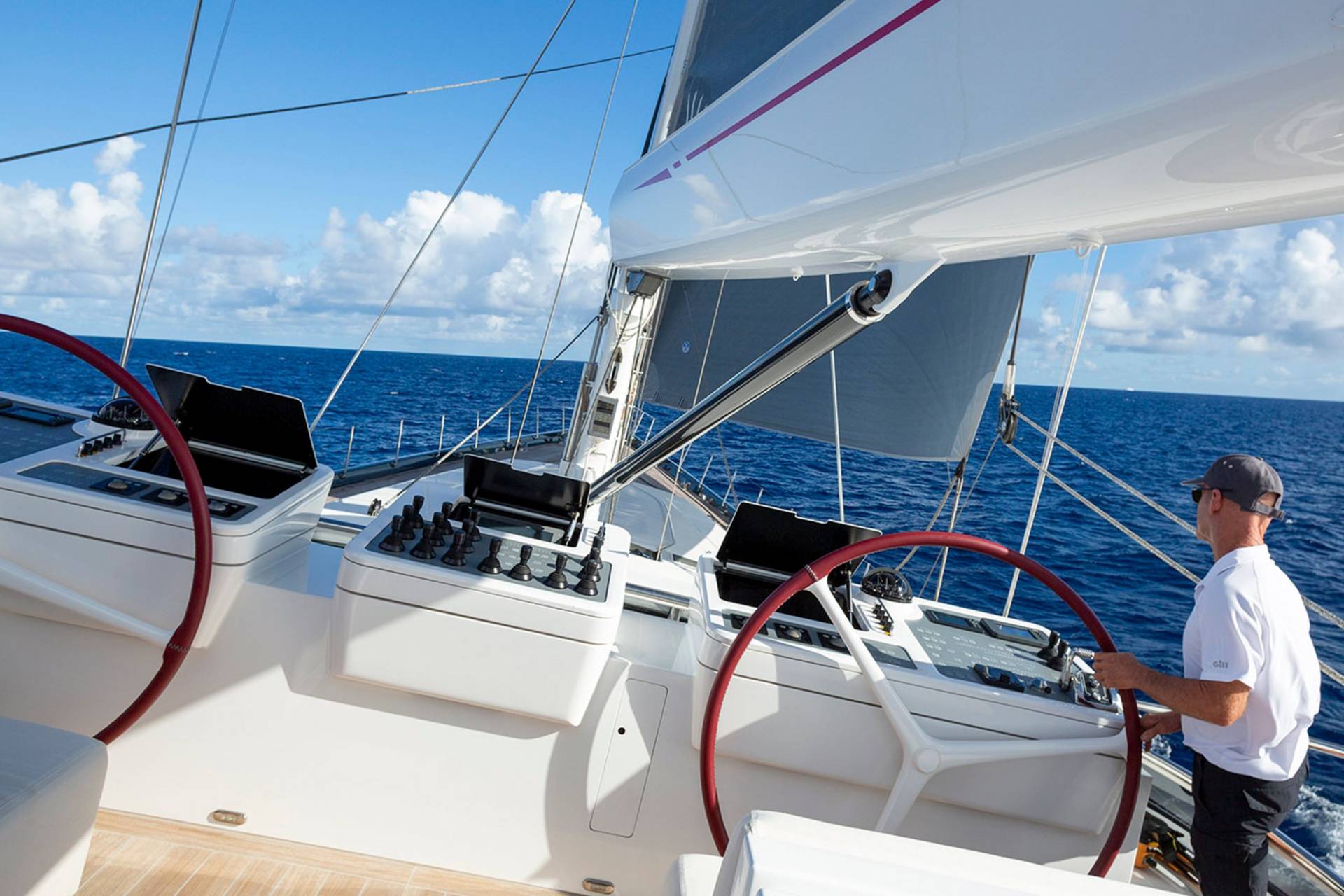 A man in a cap steers a large sailboat through the blue ocean under a bright sky.