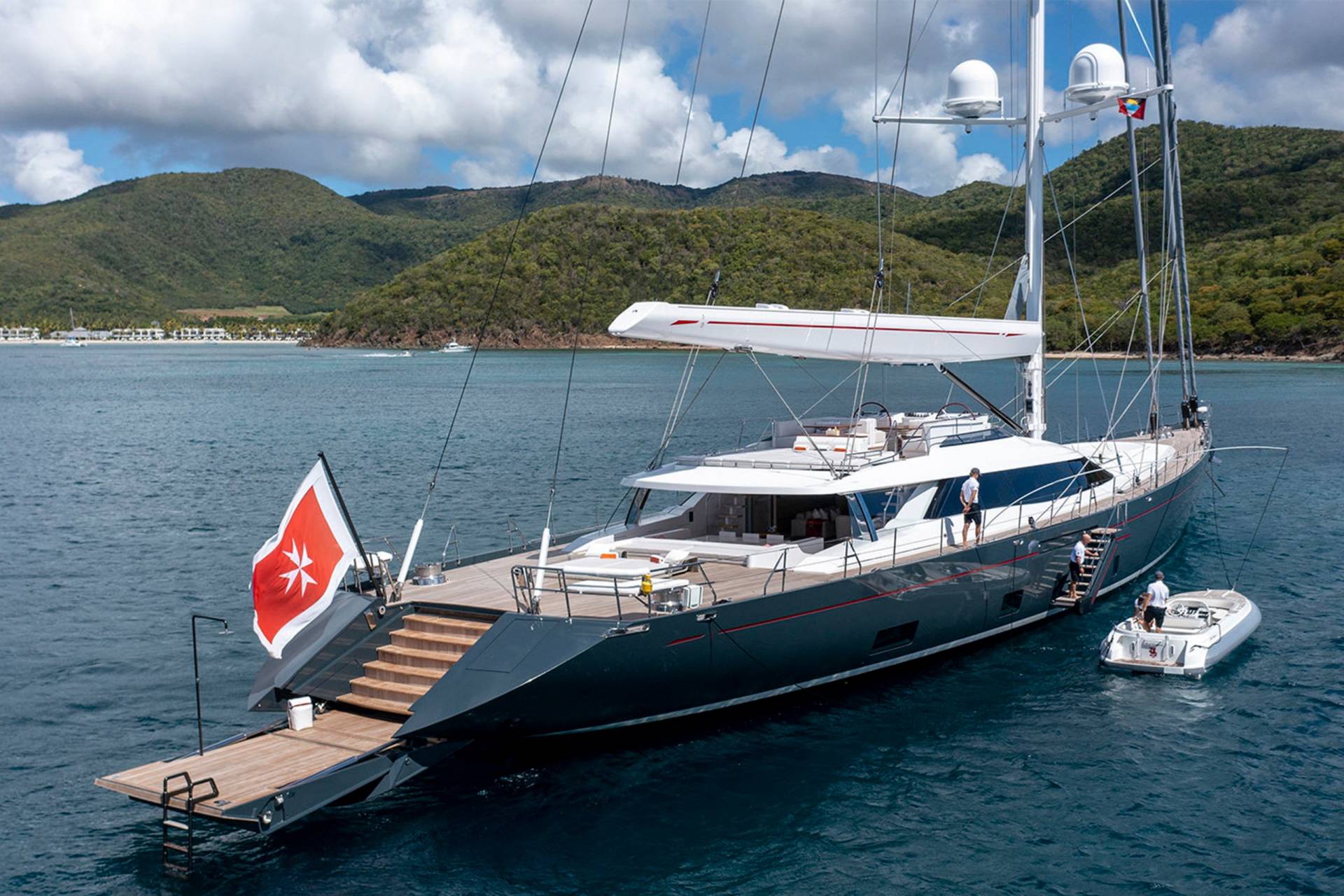 A luxurious dark grey yacht with a Maltese flag at the stern anchors in clear blue waters near a lush green coastline.
