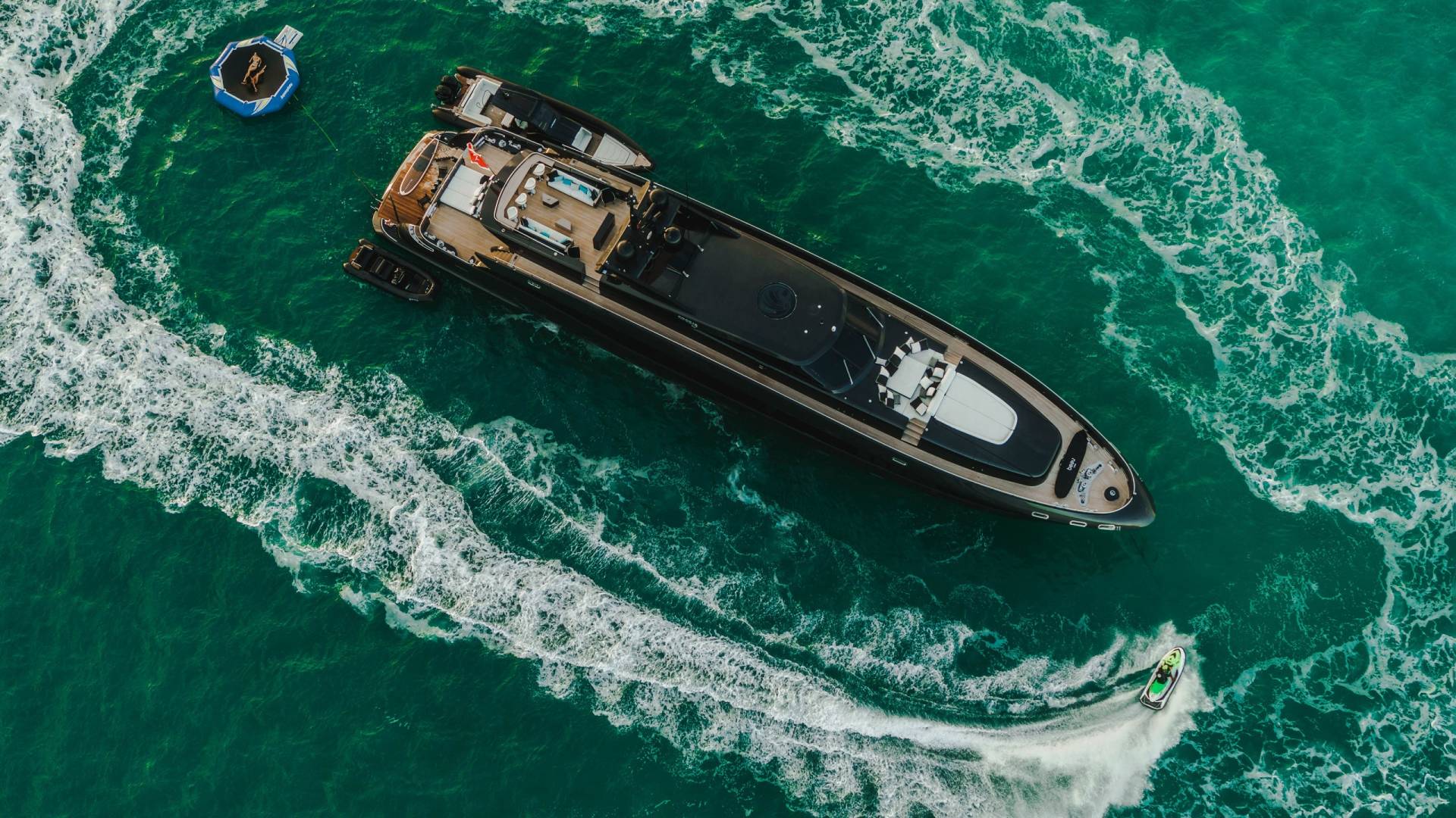 A sleek black yacht cruises through turquoise waters, trailed by a jet ski and accompanied by smaller dinghies and an inflatable water toy.
