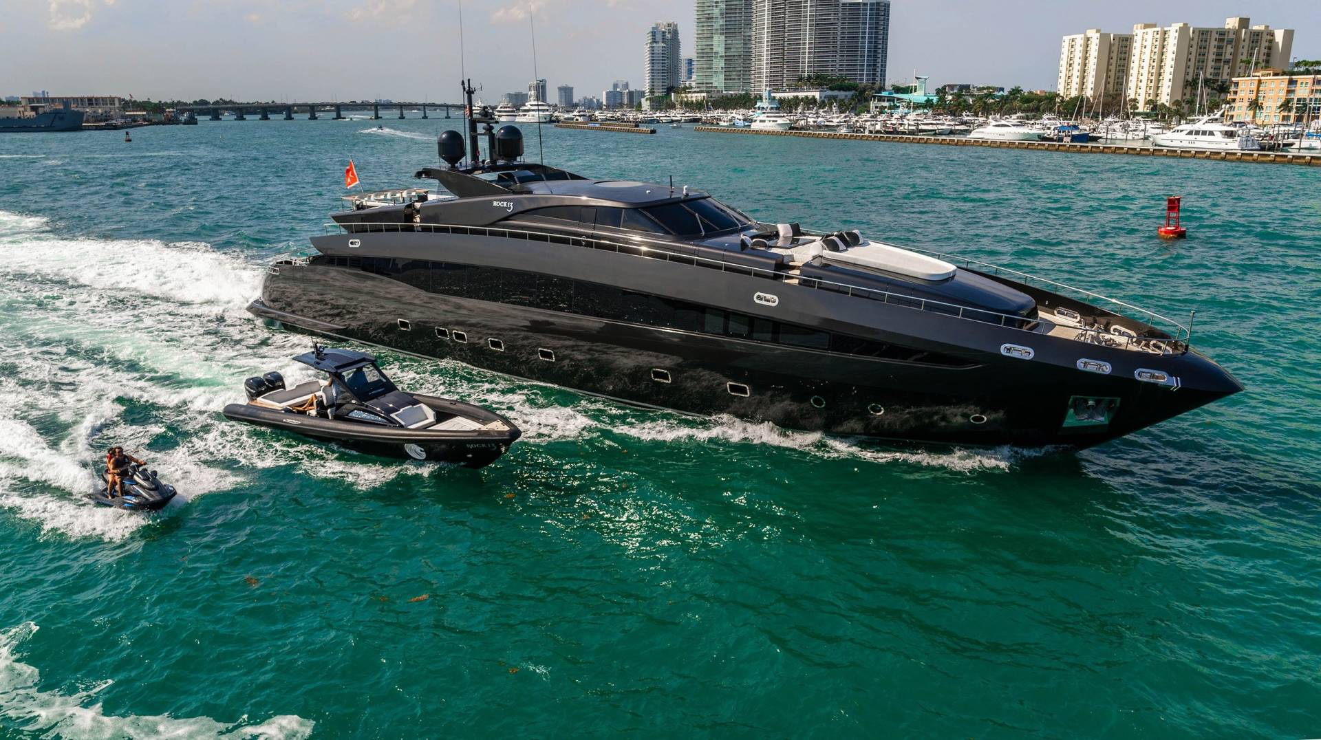 A sleek black yacht cruises through turquoise waters, trailed by a jet ski and a tender boat.