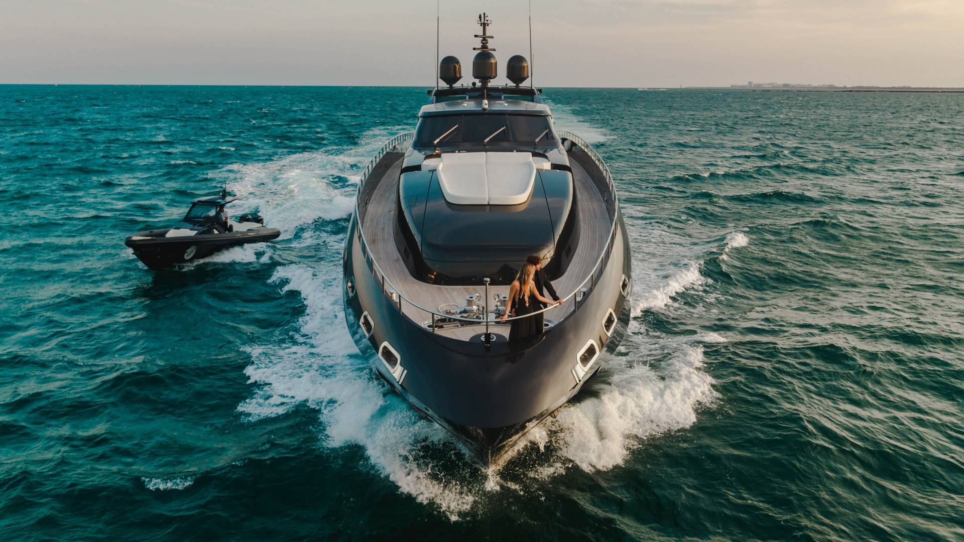 A sleek, dark yacht cuts through turquoise waters, accompanied by a smaller tender, as a couple enjoys the open sea.
