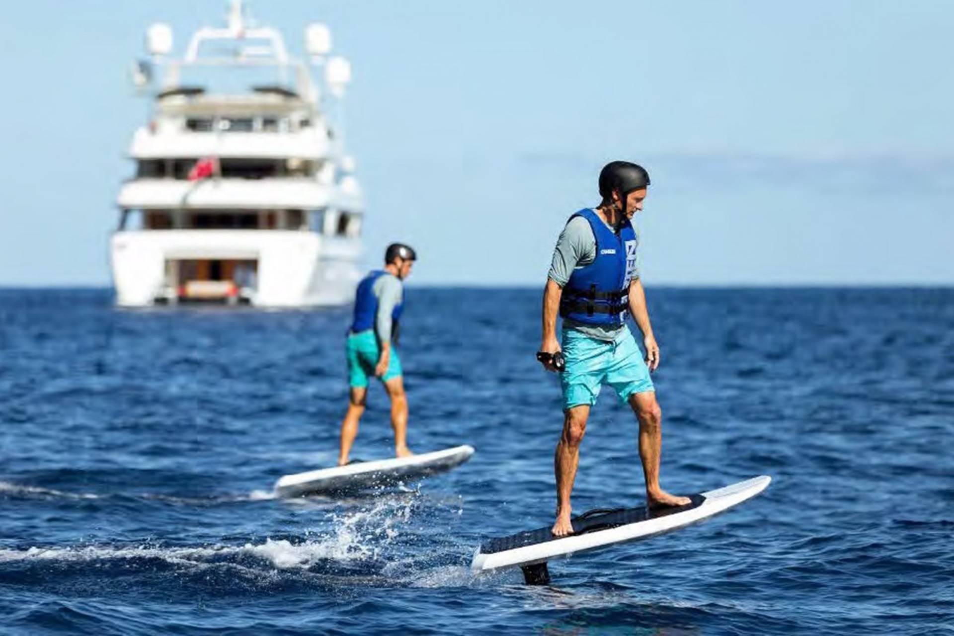 Two men on jet-powered surfboards glide across the ocean's surface with a large yacht in the background.