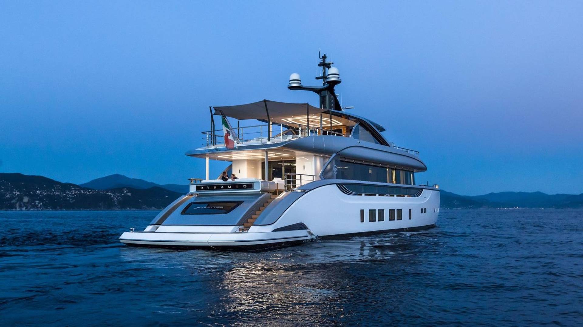 A luxurious yacht named "Dynamiq" glides through calm waters at dusk, with distant hills silhouetted against the twilight sky.