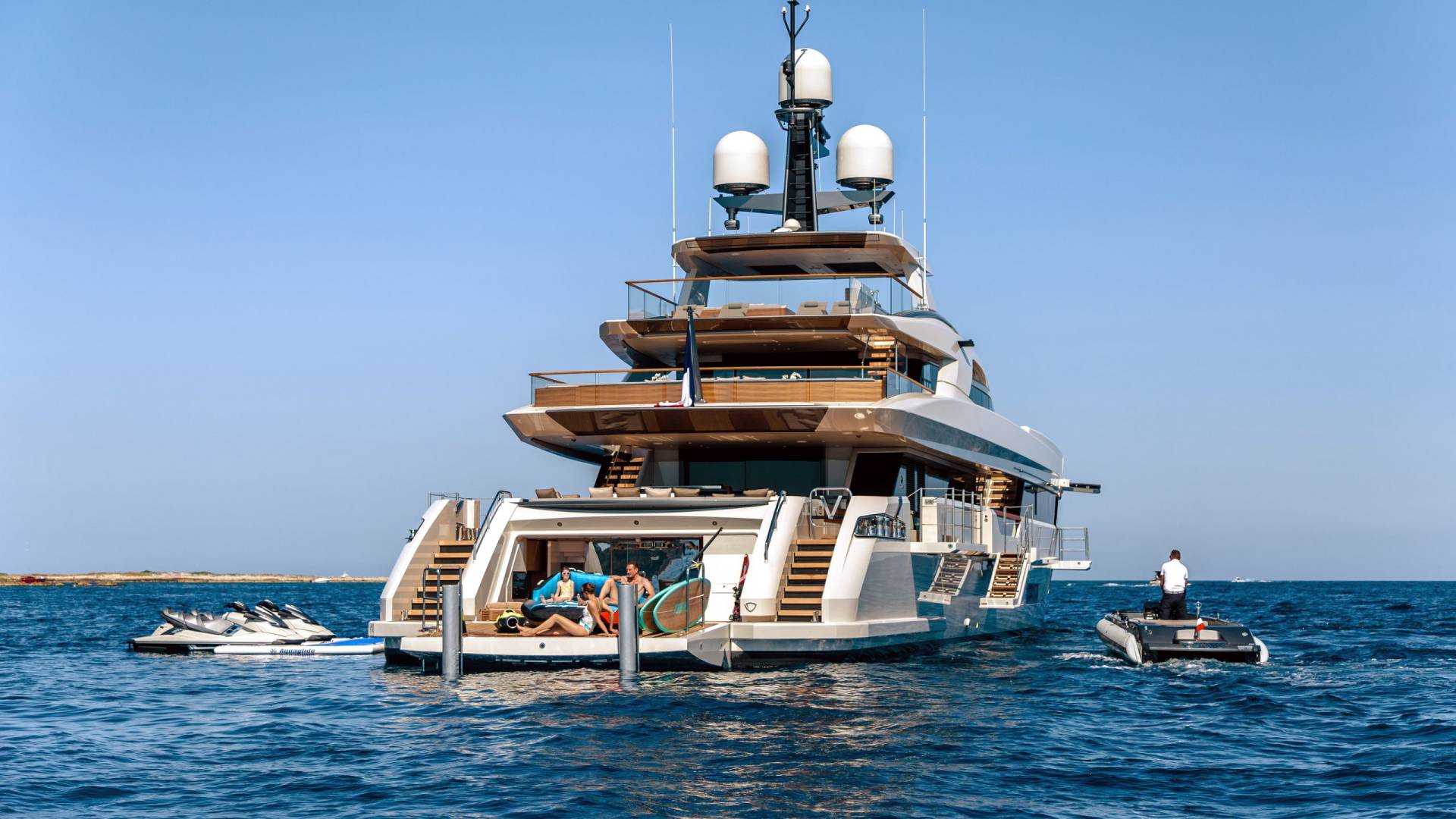 A luxurious yacht with jet skis and paddleboards awaits a group of people enjoying a sunny day on the water.