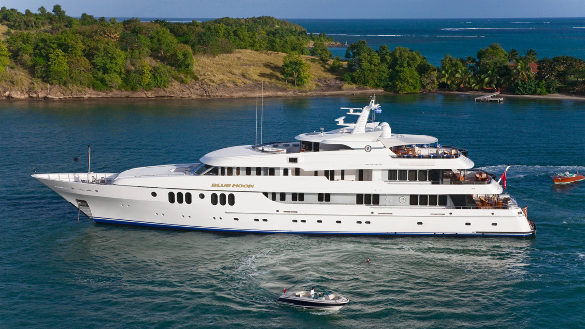 A luxurious white yacht named "Blue Moon" cruises through calm turquoise waters near a lush, green island.