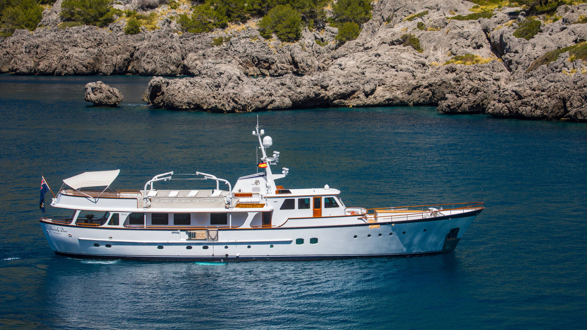 A classic white yacht cruises through the deep blue waters near a rocky, tree-lined coast.
